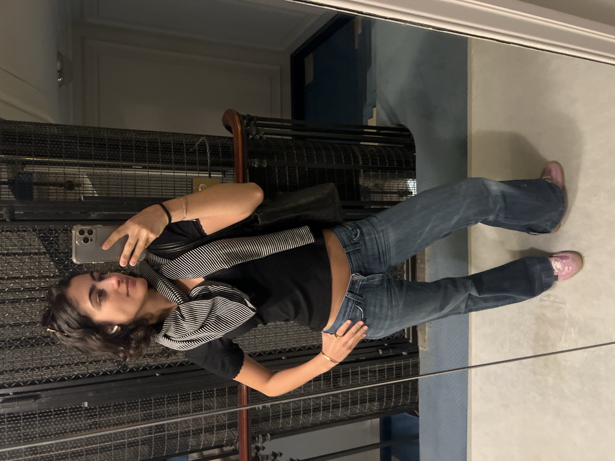 This full-body mirror selfie captures a young woman in what appears to be an older indoor setting, potentially an elevator or hallway within a building in Paris, France. She holds a smartphone with a clear case in her left hand, taking the photograph, which partially obscures her face. She has dark, curly hair, wears what look like wireless earbuds, and has sunglasses pushed up on her head. Her expression is calm and neutral.
She is dressed casually in a black short-sleeved top, blue flared jeans, and light pink sneakers. A black and white striped scarf is draped around her neck and shoulders. She accessorizes with a thin gold-colored bracelet on her right wrist and a dark band on her left. A small black bag is visible, slung over her left shoulder.
The background, reflected in the mirror, is dimly lit. A prominent feature is a large metal grated structure, reminiscent of an old-fashioned elevator shaft or decorative barrier. Behind this, parts of a wooden railing or banister are visible. The floor is a light, neutral tone, and a section of a dark blue wall or floor is reflected in the distance. The subdued, artificial lighting suggests an evening or nighttime setting indoors. No visible text is present in the image.