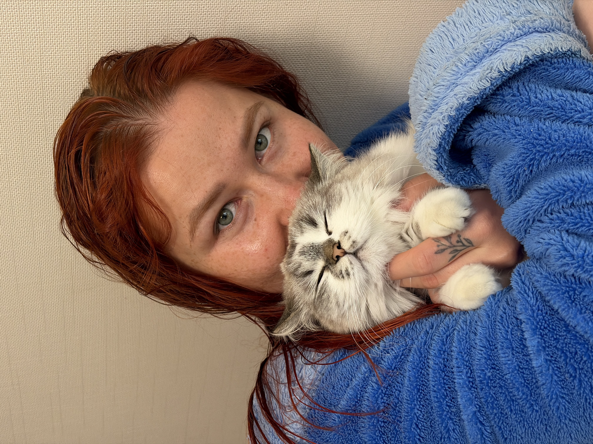 A woman with wet, red hair and green eyes is holding a light gray and white kitten in a blue bathrobe. The kitten is nestled against her cheek and appears to be sleeping. A tattoo is visible on the woman's hand. The background is a plain, neutral-colored wall. The location is not specified in the image and cannot be determined from the available information.
