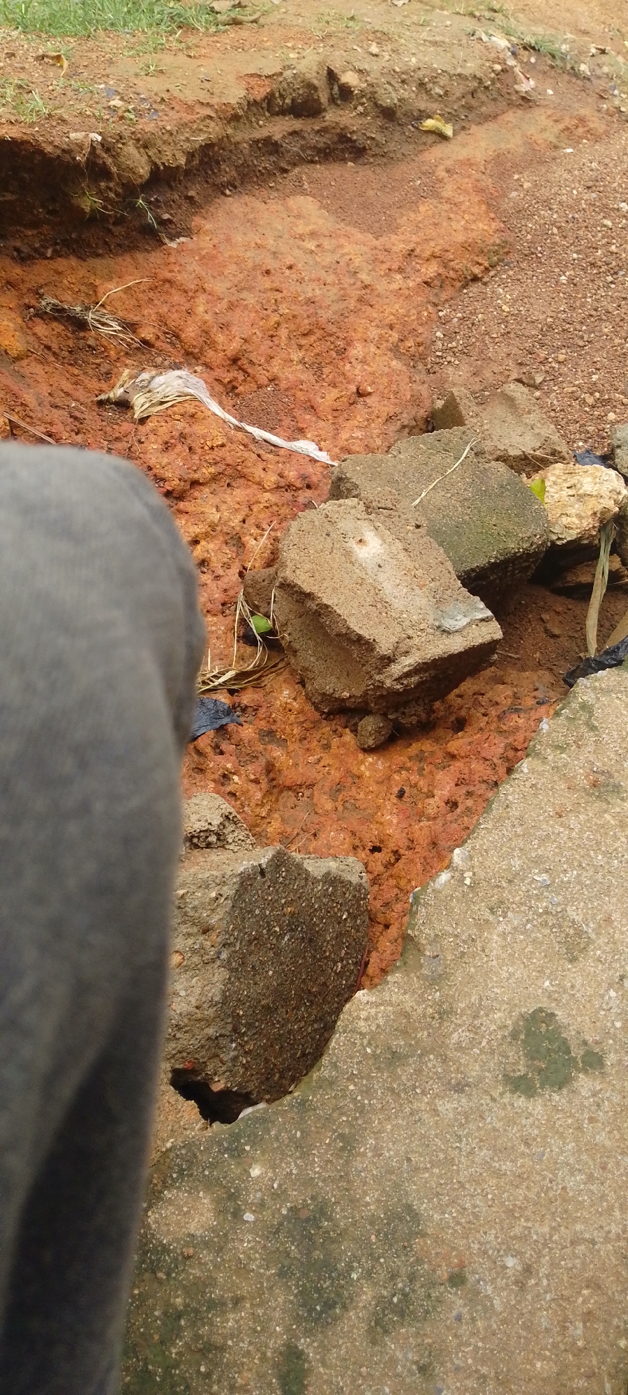 The image depicts a close-up, ground-level view of a muddy, eroded ditch in a rural or undeveloped area, possibly Baissa, Nigeria. The ditch is characterized by its prominent reddish-brown, clay-like soil, suggesting a mineral-rich composition or perhaps iron content. Several rough, grey concrete blocks are scattered within and around the ditch, some appearing broken or weathered. Among the debris, a piece of white plastic sheeting is visible, indicating litter or construction remnants. The surrounding ground is a mix of soil, gravel, and sparse green grass.
In the foreground, a large portion of a person's leg, covered by dark grey fabric, is visible on the left, obscuring part of the scene and suggesting the photographer's perspective. The lighting suggests it is daytime, with diffused light, possibly indicating an overcast sky or shaded conditions. There is no discernible text in the image. The overall impression is one of neglect and environmental degradation, with man-made debris interspersed with natural elements in a state of erosion.