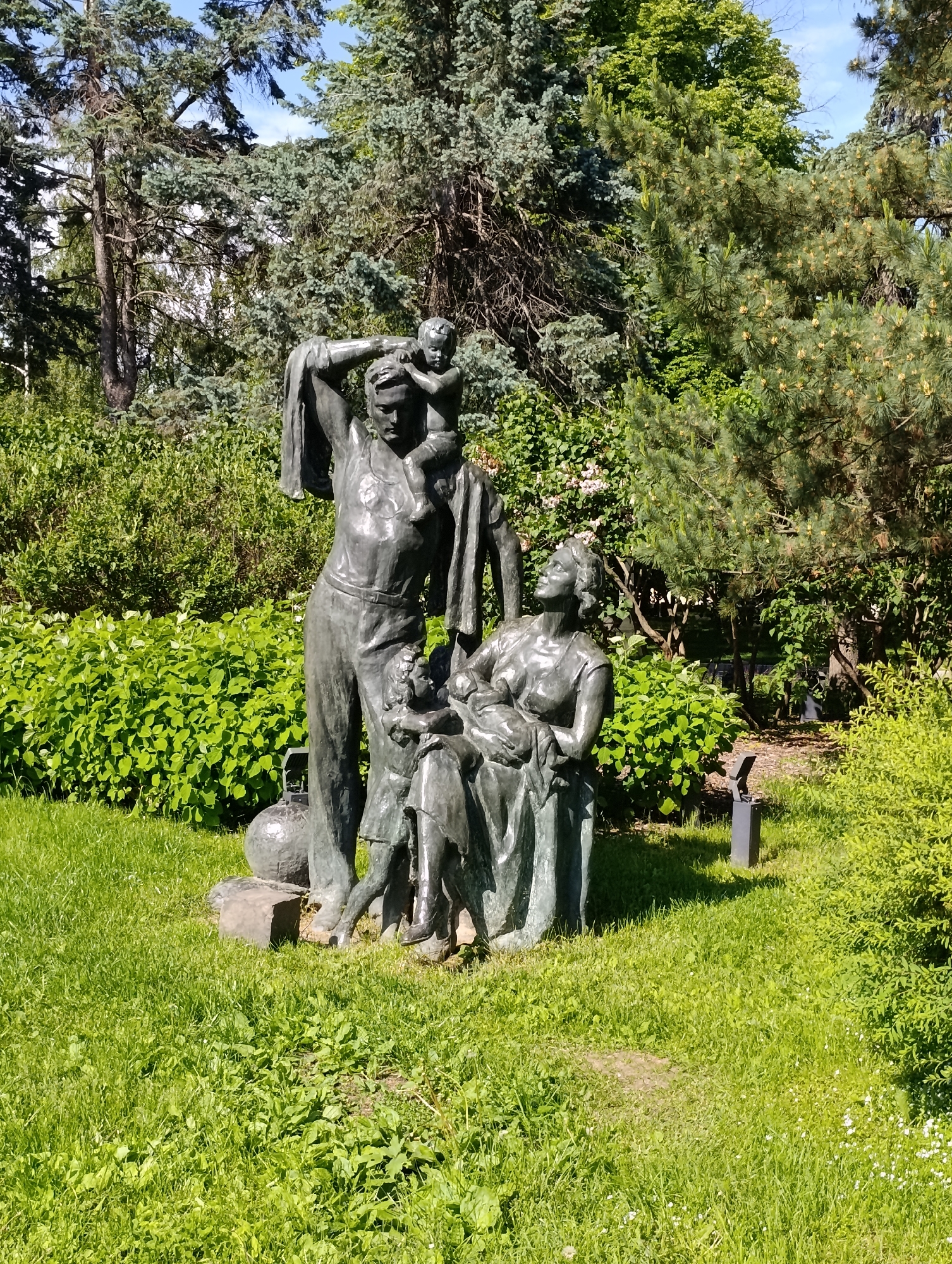 This media file captures a bronze sculptural group depicting a family, set within a lush green park in Moscow, Russia. The central subjects are four figures cast in bronze. A strong, standing man, appearing to be a worker in overalls, supports a small child on his right shoulder while holding a garment or cloth over his left shoulder. Next to him, a woman is seated, cradling an infant in her lap. Another young child stands between the man and woman, seemingly interacting with them.
The statue is positioned on a patch of vibrant green grass, which shows some wear, hinting at paths or foot traffic. Surrounding the monument are thick, leafy green bushes and a dense background of various trees, including tall conifers with blue-green needles and deciduous trees with bright green foliage. The scene is brightly lit by natural sunlight, suggesting a clear day, likely midday or early afternoon, with visible shadows cast by the figures and vegetation. A patch of clear blue sky is visible through the canopy of trees. Two small, dark, modern ground lights are partially visible embedded in the grass near the base of the statue, suggesting evening illumination for the artwork.
The activity depicted is a static, posed representation of family life and possibly labor, emphasizing unity and nurturing. The man's stance and the presence of children suggest themes of protection, provision, and continuity. No visible text is present on the statue or in the immediate surroundings. The monumental style and themes are characteristic of public art found in parks within the region.