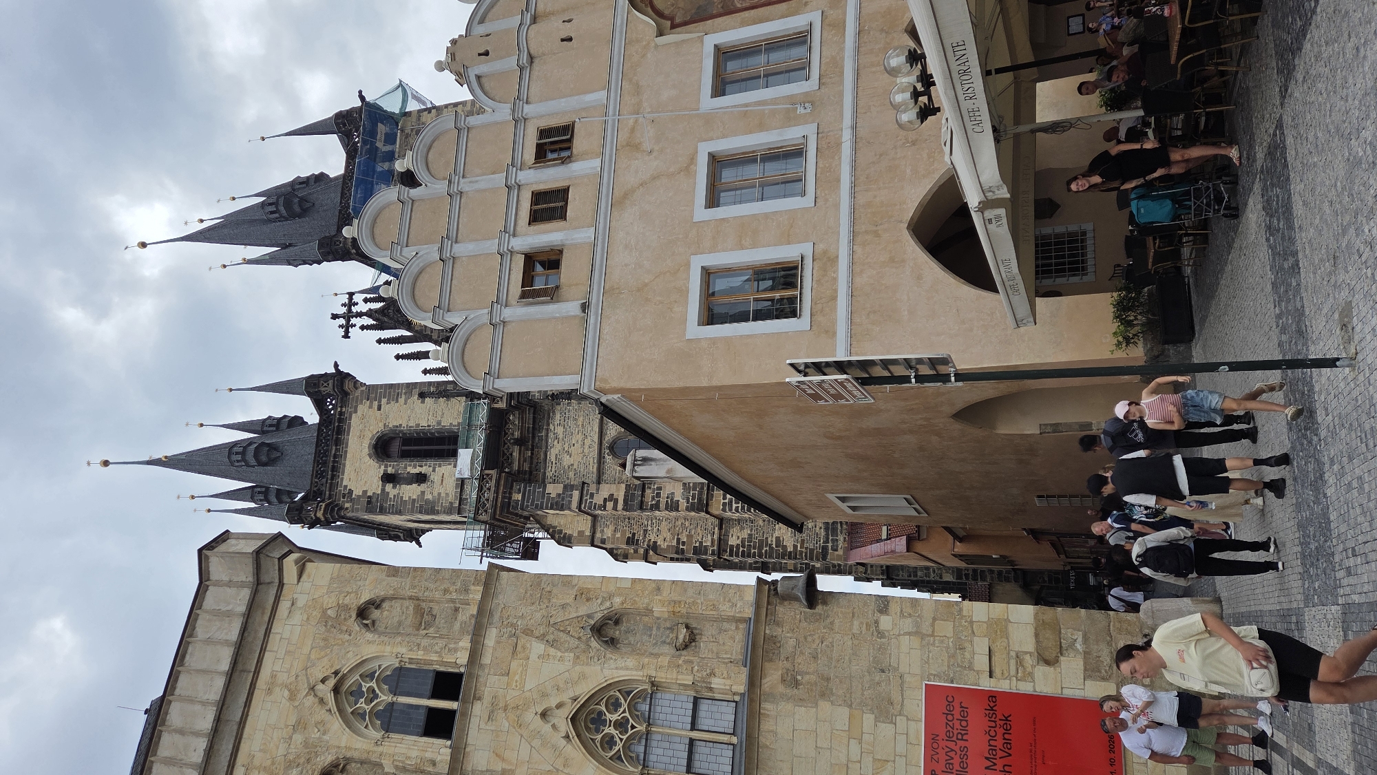 This is a street-level, eye-level, medium shot photo of the Old Town Square in Prague, Czechia. The iconic Týn Church dominates the background with its gothic spires. To the left of the Týn Church, a lighter colored building with gothic architecture stands prominently. In the foreground, people are walking on a cobblestone street. On the far left, a large red poster is affixed to a building. A cafe with outdoor seating and umbrellas is visible on the right side of the image, with "CAFFE - RISTORANTE" written on the umbrellas. The sky is overcast.