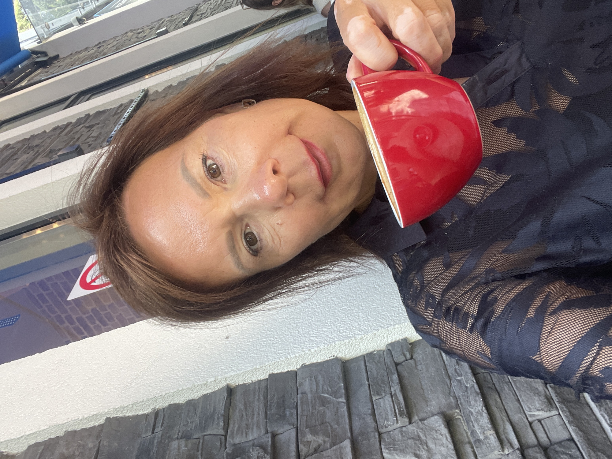 A middle-aged Caucasian woman with shoulder-length brown hair is taking a selfie in front of a stone and stucco building. She is holding a red cup of coffee in her right hand and is wearing a black lace-patterned shirt. The woman is looking at the camera with a slight smile. The background shows a window with a blue awning, and a red and white sign. The image is taken outdoors in daylight. There is no indication of the city of Sochi, Russia in the image.