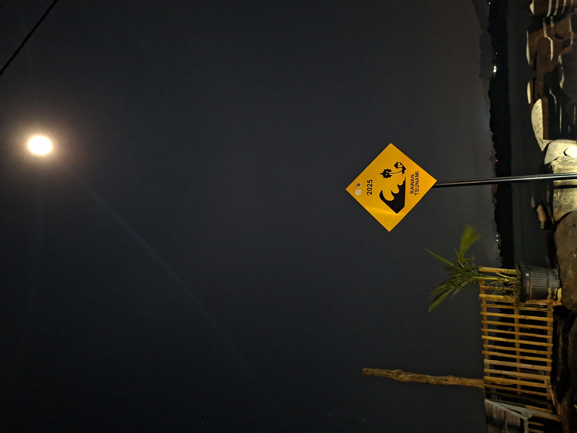 The image captures a nighttime scene with a dark sky, illuminated by a bright, spherical light source, likely the moon, in the upper left quadrant. A prominent yellow, diamond-shaped warning sign is positioned centrally to the right, mounted on a dark pole. The sign features black text and symbols: "2025" is written vertically on the left side, and on the right, the words "RAWAN TSUNAMI" (Indonesian for "Tsunami Prone" or "Tsunami Hazard") are displayed, accompanied by a graphic depicting a large wave, a person running, and a palm tree.
Below the sign, a small palm tree in a dark pot is visible, resting on a wooden slatted structure, possibly a railing or a small deck. To the right and extending downwards, a darker, rough-textured structure can be seen, which appears to be part of a larger wooden or concrete embankment or foundation. In the background, to the right of the sign and structure, there is a dark expanse that suggests a body of water, possibly the sea or ocean, with faint reflections of distant lights on its surface. A dark wire or cable runs diagonally from the upper left corner of the frame. The overall impression is a nocturnal coastal area in Lhokseumawe, Indonesia, marked by a tsunami warning sign, indicating preparedness or awareness for potential natural disasters.
