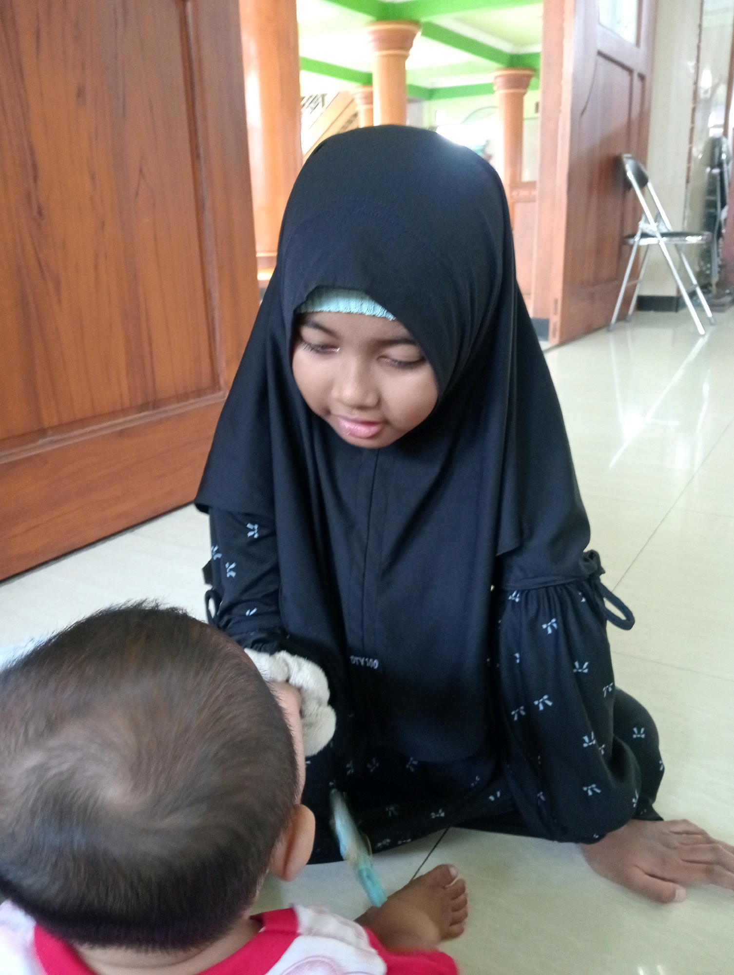 Two young children, one wearing a black hijab and the other a red and white shirt, are interacting in a room. The child in the hijab is seated and looking down, while the other child, whose back is to the camera, is also seated on the floor. The environment appears to be indoors, with wooden doors and tiled flooring visible. The room also contains pillars and a chair in the background, suggesting it might be a community space or a home. The overall scene depicts a moment of interaction between the two children.