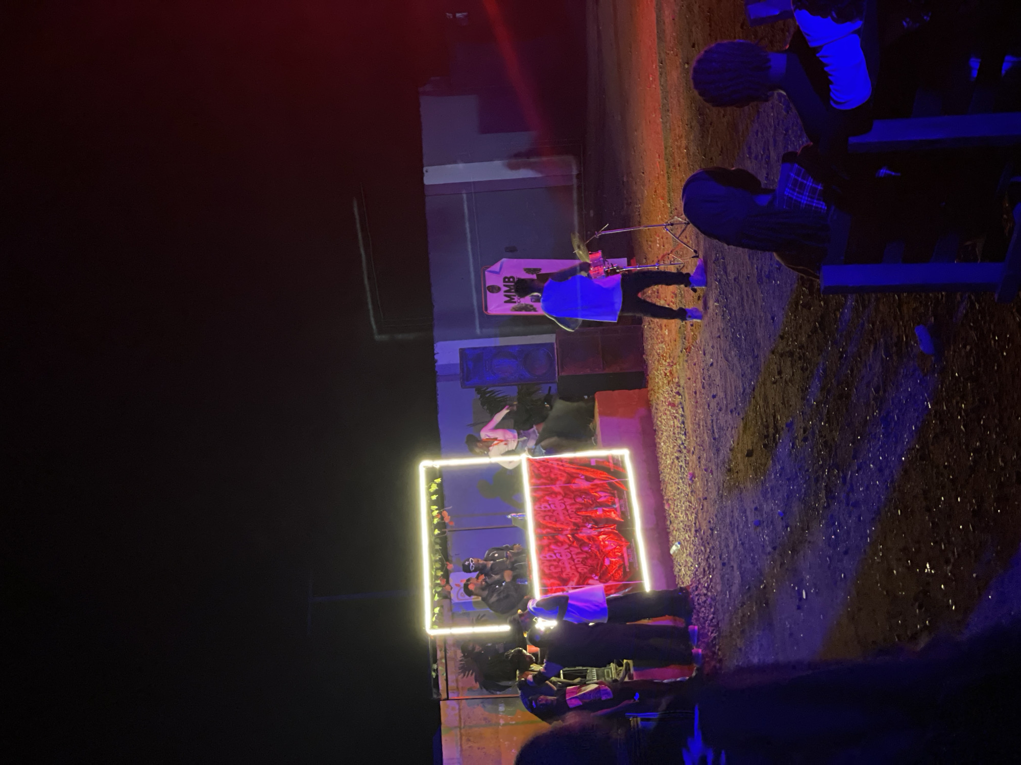 An outdoor event is taking place at night in Durumi, Nigeria. A brightly lit DJ booth, outlined in neon lights, is central to the scene. Two individuals are behind the DJ booth, appearing to be performing. A banner with red and black graphics that reads "MMB BACK PARTY" is draped across the front of the booth. To the right, a person is playing drums. In the foreground, several people are seated on benches, facing the performance. The ground is covered in gravel, and the background features a building structure with some lighting. The atmosphere suggests a lively gathering or concert.