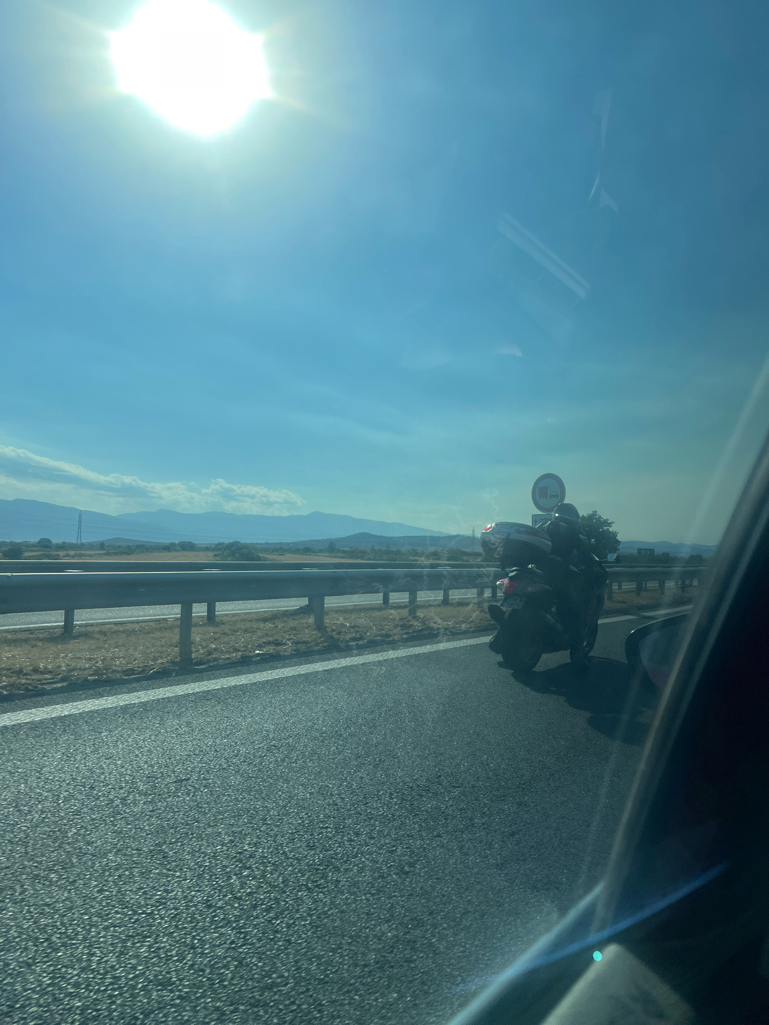 A scooter is on a highway, seen from a car. The scooter is dark, with a large storage box on the back. A rider wearing a helmet is on the scooter. There is a road sign behind the scooter that appears to be a prohibition sign for trucks. The highway has a metal guardrail on the left side. In the background, there are mountains and a bright sun in the sky. The image appears to be taken in a rural area.