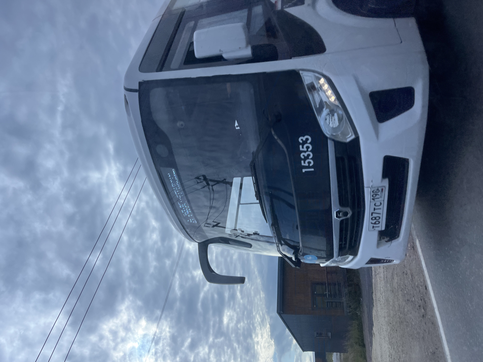 The image shows the front of a white bus with a black front panel. The bus is positioned on a paved road, with a dirt shoulder to its left. Behind the bus, there is a long, low-slung building with a corrugated metal exterior. The sky above is overcast with dramatic, cloudy formations. Power lines are visible in the upper portion of the image. The bus has a license plate that reads "T687TC 196". On the front of the bus, the number "15353" is displayed prominently in white text. The image appears to be taken in Glyadino, Russia, based on the license plate format, which is typical for Russian vehicles. There are no people visible in the image.