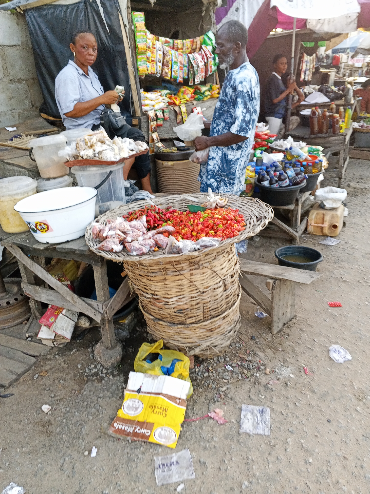 The image depicts a bustling market scene in Zango-ndaji, Nigeria. A woman sits in front of a stall counting money. A man wearing a blue and white patterned shirt stands at a stall holding a plastic bag. In front of them is a large woven basket filled with what appears to be peppers and bags of unidentified items. Several other stalls are visible in the background, displaying a variety of goods, including packaged foods and bottled drinks. The ground is littered with debris, including a flattened yellow box with "Curry Masala" printed on it.