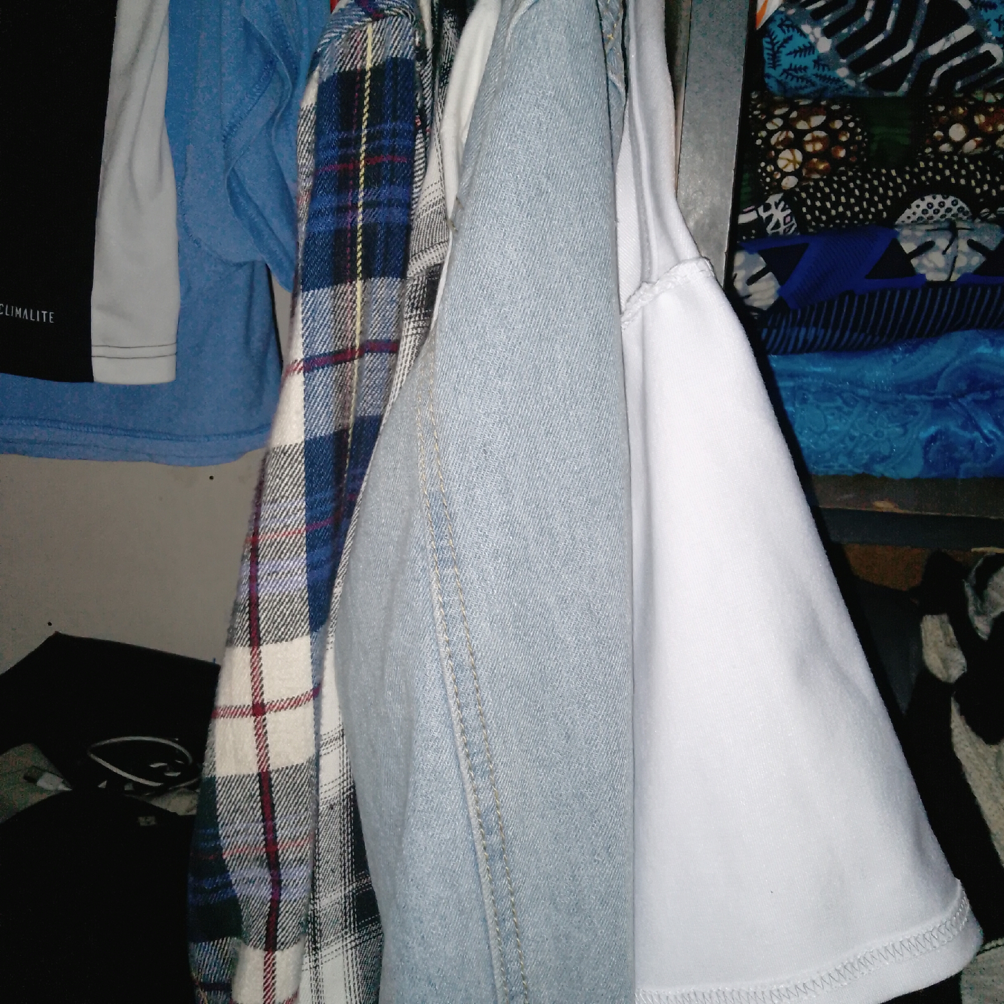 The image displays a close-up view of several items of clothing hanging together. The primary subjects are a black t-shirt with "CLIMALITE" text visible, a blue long-sleeved shirt, a plaid shirt in shades of blue, white, and red, and a white t-shirt. There are also glimpses of patterned fabrics stacked in the background. The setting appears to be a closet or wardrobe, with the clothes hanging on what looks like a metal rod. The lighting is dim, suggesting an indoor environment, possibly at night or in a poorly lit room. No specific activity or event is depicted, other than the clothes being stored. The focus is on the textures and patterns of the garments, with the stitching on the white t-shirt and the seams on the blue denim-like garment being subtly visible. There are no discernible emotions conveyed by the image, as it is a still life of inanimate objects. The time of day and weather are not indicated. The location context is unknown.