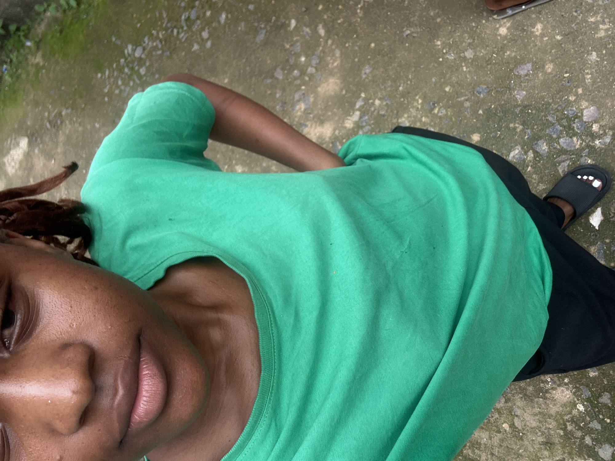 This media file is a close-up, high-angle shot of a person wearing a bright green t-shirt and black pants, standing on an outdoor paved surface. The person's face is partially visible in the foreground, showing their lips and nose. Their left arm is bent, with their hand resting on their hip. Their right leg is visible in the lower right of the frame, wearing a black sandal with white painted toenails. The ground is a textured, grey concrete surface with small stones and patches of moss. In the background, there are hints of green foliage. The lighting suggests it is daytime. The overall mood appears casual and relaxed. There is no visible text in the image. The context suggests this image was taken in Rafin Par, Nigeria.