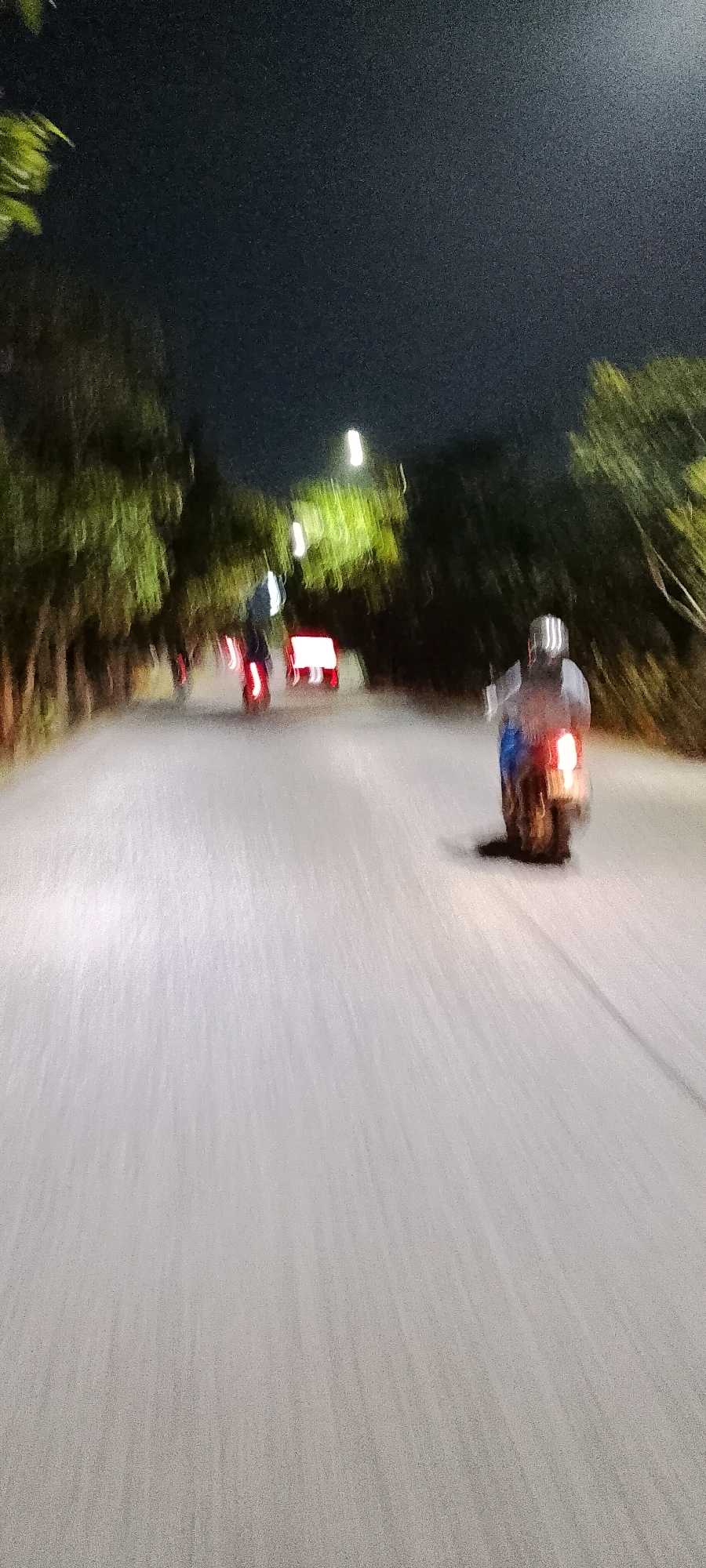 The image depicts a nighttime scene on a road in Bekasi, Indonesia, characterized by motion blur. Two individuals are visible riding motorbikes. The motorbike on the right is closer to the foreground and its rider is wearing a helmet. The motorbike on the left is further back and its rider is also wearing a helmet. Both motorbikes have illuminated taillights, indicated by red streaks of light. The road surface is light-colored and appears wet or reflective. Trees line both sides of the road, with some artificial lights visible among them, casting a green glow. The sky above is dark. The overall impression is of a dynamic and somewhat indistinct urban environment at night.