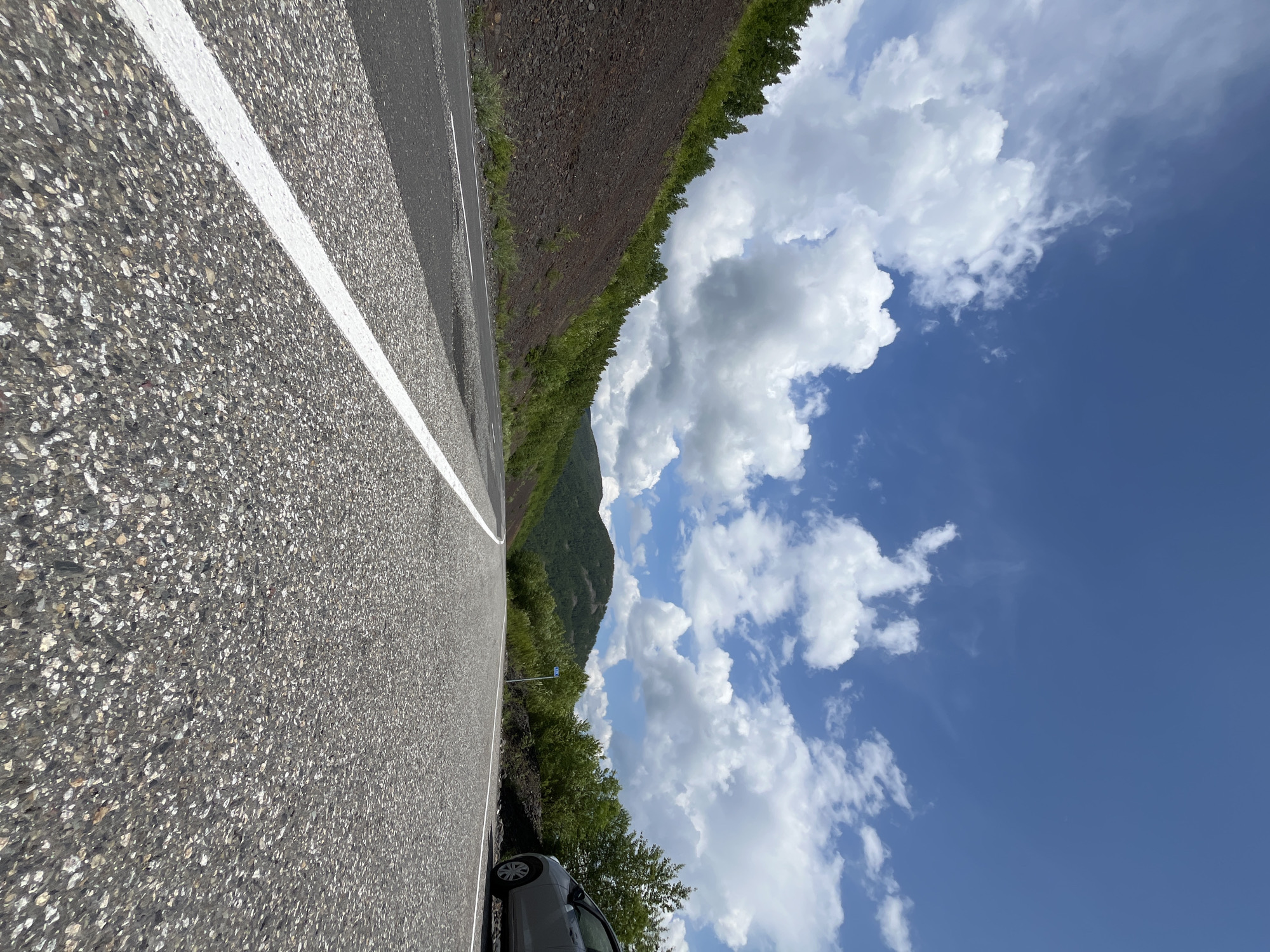 A low-angle, eye-level shot captures an asphalt road in Russia, winding through a landscape of rolling green hills and rocky slopes under a bright, partly cloudy blue sky. The road, marked with white lines, curves to the left and disappears into the distance. To the left, a steep, dark, rocky slope is covered in sparse green vegetation. To the right, the road is bordered by trees and a silver car parked at the shoulder. The sky is filled with large, fluffy white clouds against a deep blue expanse.