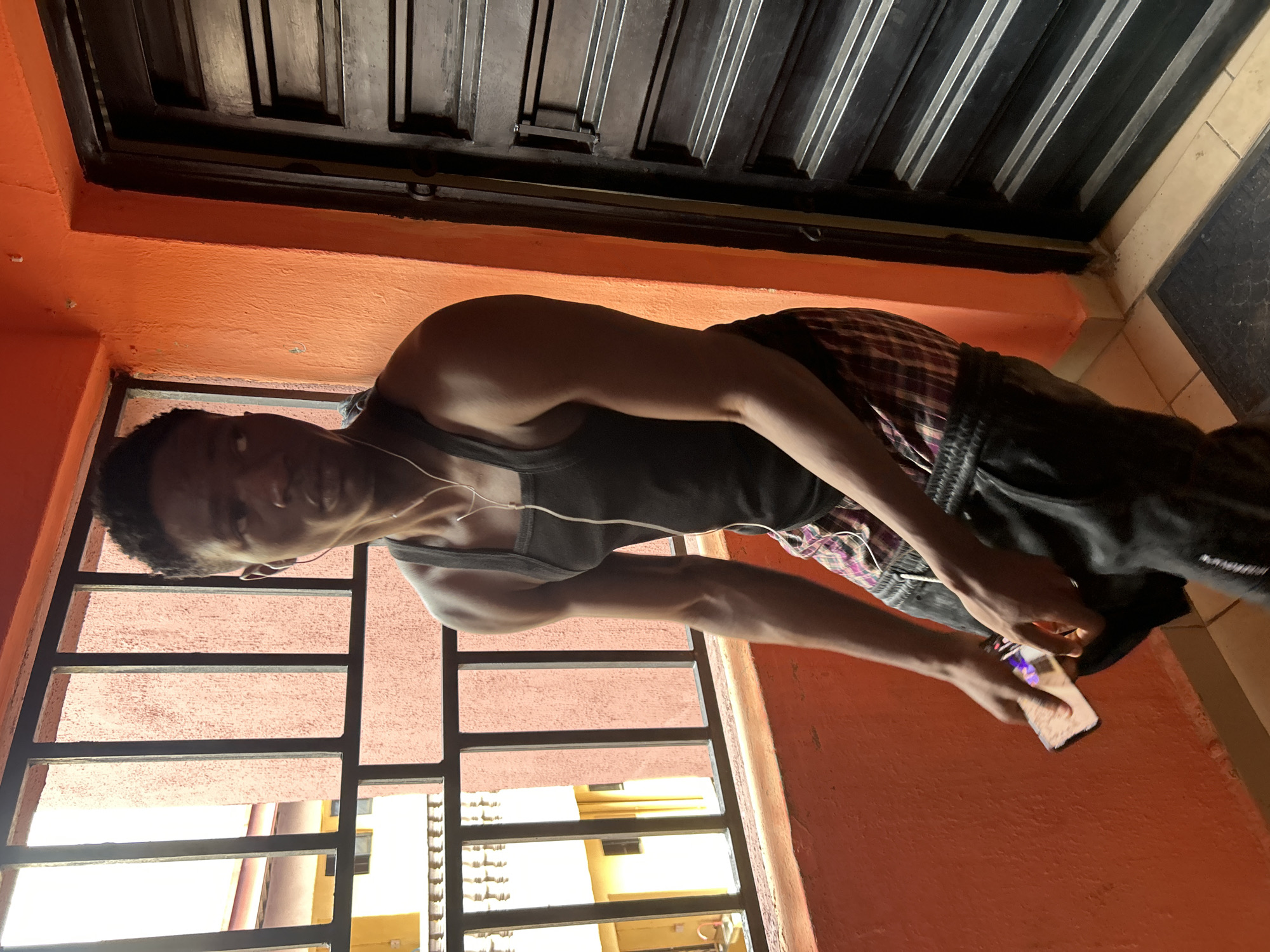 A young Black man with short, dark hair stands on a balcony in what appears to be a residential building in Ihiagwa, Nigeria. He is wearing a black tank top, plaid shorts with a red, purple, and white pattern, and dark sweatpants. He has white earbuds in his ears and is holding a smartphone in his right hand. The background shows a metal railing with vertical bars, beyond which are the facades of other buildings. The wall on the right is painted bright orange, and a dark metal door or gate is visible. The floor is tiled. The lighting suggests it is daytime, with natural light coming from the left, creating shadows.