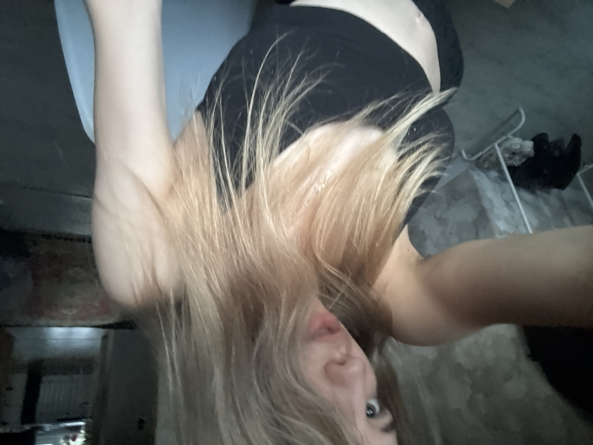 This inverted selfie features a person, likely female, with blonde or light brown hair flowing downwards due to gravity, partially obscuring their face. The individual wears a black top, possibly a sports bra, revealing part of their midriff. One arm is visible, extended upwards in the inverted frame, resting against a light-colored surface that could be bedding or furniture.
Through the strands of hair, one light-colored eye and a portion of the nose and slightly parted lips are discernible, with the gaze directed towards the camera. The overall expression is casual, possibly playful given the unusual pose.
The background depicts a dimly lit indoor setting, consistent with a room in Noviy, Russia. To the right, a white structure resembling a clothes rack or shelving unit holds some dark items. The room appears to have simple, unadorned walls. The low lighting suggests an indoor environment lacking strong illumination or an evening scene. No visible text is present.