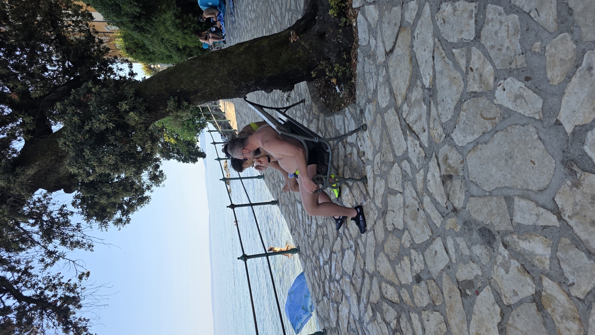 A man and a boy are sitting on a bench by the sea. The man is wearing black swim trunks and black water shoes. The boy is wearing yellow swim trunks. They are both looking out at the sea. There is a blue umbrella in the water. The ground is paved with stones. There is a large tree to the right of the bench. In the background, there are other people sitting on the beach and a building. The sky is clear and blue.