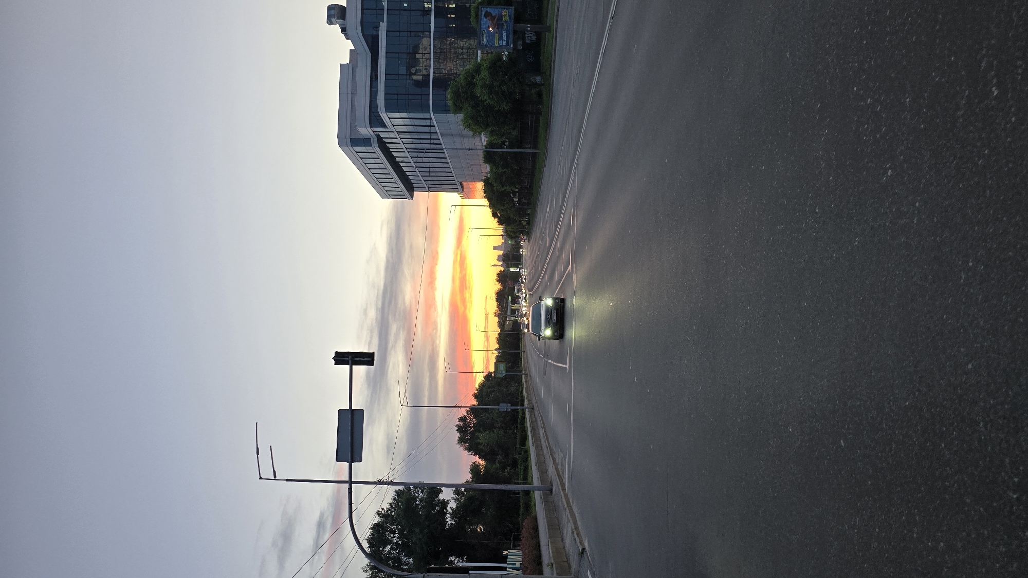 The image depicts a street scene in Kyiv, Ukraine, during a sunset. A modern, multi-story building with a glass facade dominates the right side of the frame. A dark-colored car is driving on the asphalt road in the foreground, with its headlights on. Several other cars are visible further down the road, their lights creating streaks against the twilight sky. The sky itself displays a gradient of colors, with orange and yellow hues near the horizon, transitioning into pale blue and gray. Trees line the left side of the road, and a traffic light is visible in the upper left quadrant. The overall impression is one of urban twilight, with a blend of modern architecture and natural light.