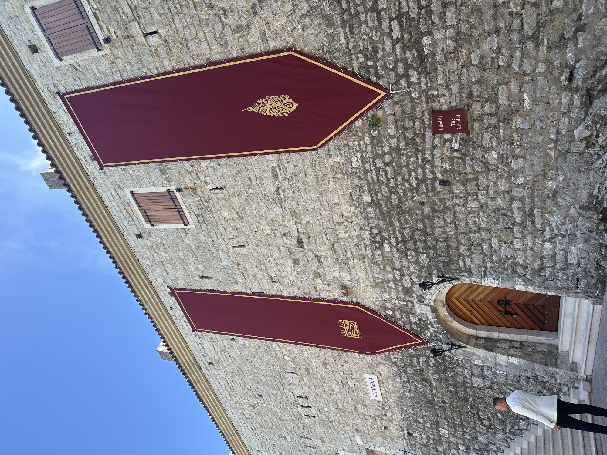 Here is a detailed description of the image:
The image depicts a stone building, likely a historic structure, with two large burgundy banners hanging on its exterior. Each banner has a gold border and a unique emblem: the upper banner features an intricate gold crest, while the lower banner displays the word "BUDVA" above another design. The building's windows have wooden shutters.
A sign reading "Citadela / The Citadel" is fixed to the wall. A wooden door is set into the stone wall, accessed by a set of stairs. A person is partially visible at the bottom of the frame, wearing a white jacket and dark pants, ascending the stairs toward the door. The sky is visible in the upper left corner of the frame and appears blue.
This scene is located in Budva, Montenegro, as indicated by the "BUDVA" banner and the "Citadela" sign. The image captures a moment of potential pedestrian interaction with a historic site in the city.