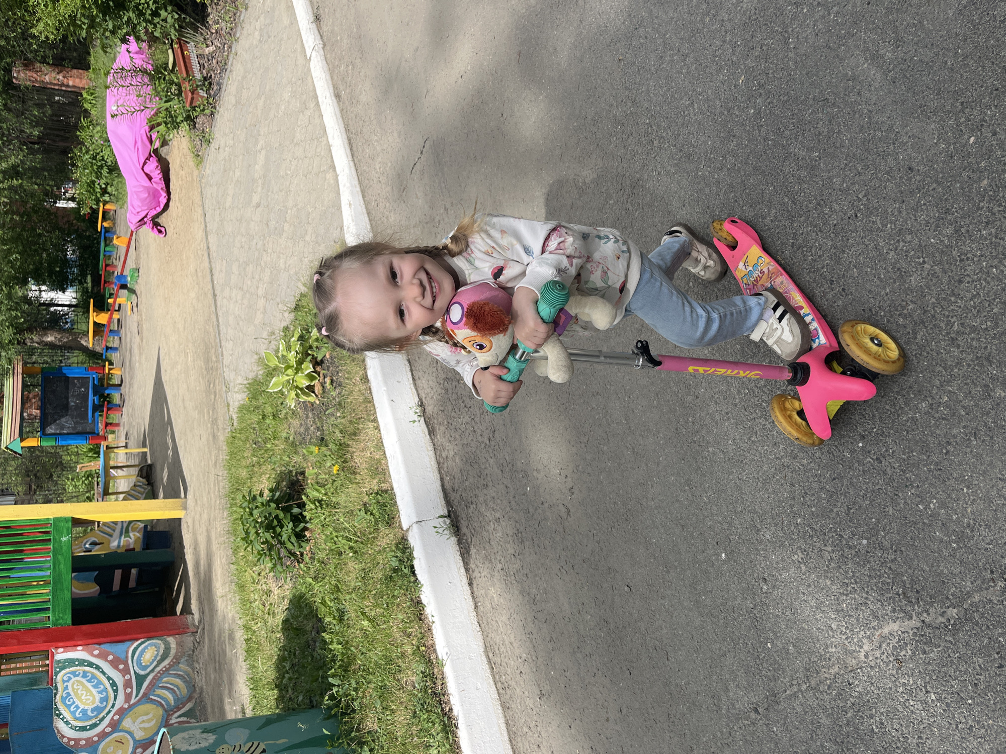 A young girl is captured in an outdoor setting, likely a playground or park, on a bright, sunny day. She is smiling broadly at the camera, showing a cheerful and happy disposition. Her light-colored hair is styled in two pigtails, secured with pink ties. She is wearing a long-sleeved white top with a subtle floral pattern, light blue jeans, and light-colored sneakers with dark stripes.
The girl is positioned on a pink kick scooter with yellow wheels. The brand name "SKECHERS" is visible on the scooter's stem, and the word "GOOD" can be seen on the deck. She is holding onto the scooter's handlebars with one hand, while cradling two stuffed animals and a green toy in her arms. One stuffed animal is pink and brown, possibly a dog or monkey, and the other is lighter in color.
The setting includes a paved asphalt surface, a concrete sidewalk or path, and strips of green grass and foliage. In the background, there is colorful playground equipment, including a wooden structure painted in vibrant blues, greens, yellows, and reds. A long, bright pink fabric tunnel or slide cover is also visible among the greenery. The sun casts distinct shadows, indicating midday conditions.