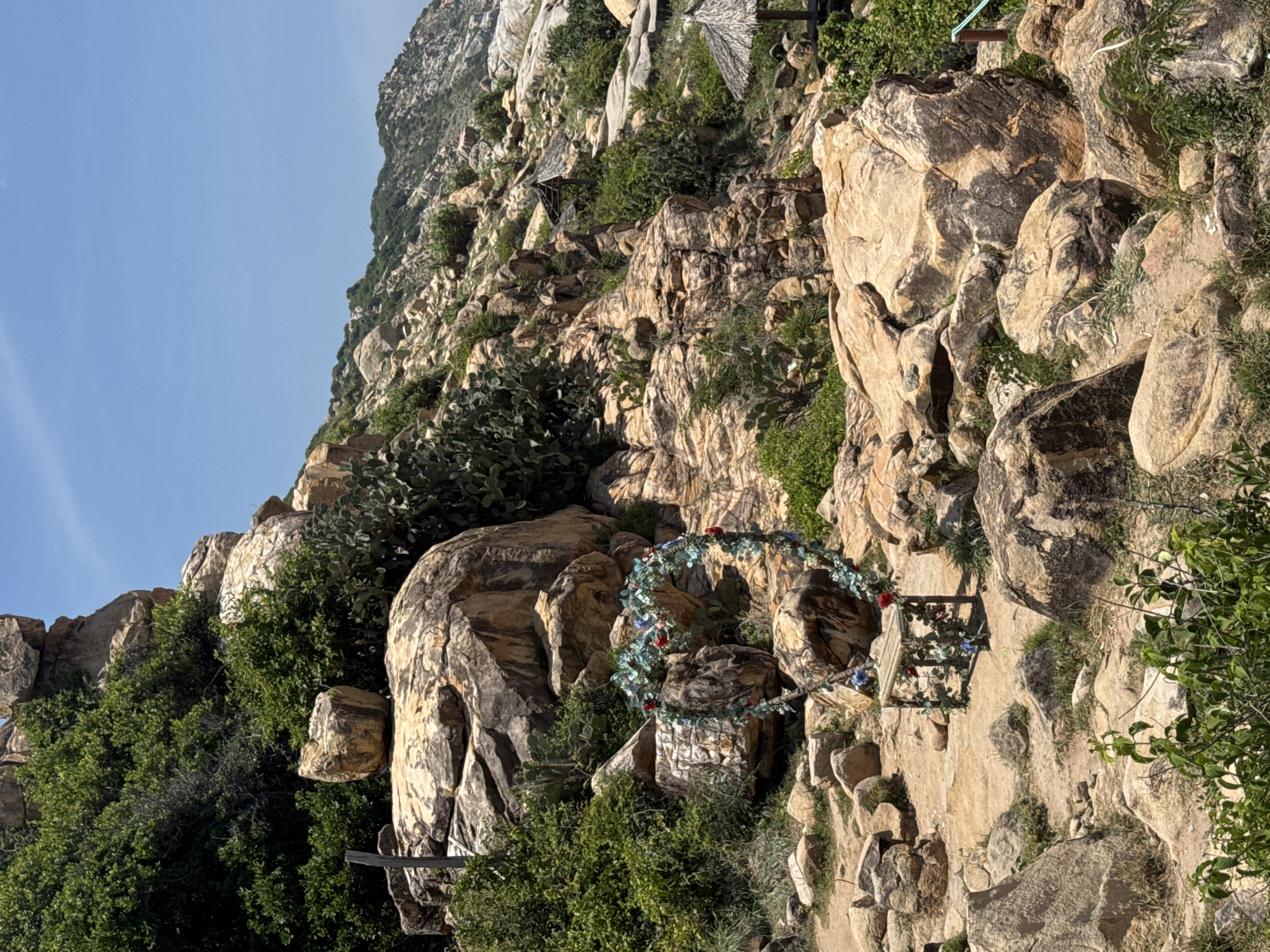 In Vinh Hai, Vietnam, a heart-shaped floral archway adorns a rocky, arid landscape. The archway, crafted from intertwined greenery and red and blue flowers, is set against a backdrop of large, weathered rocks and sparse vegetation. A small, rustic wooden table sits beneath the arch, also decorated with similar floral elements. In the distance, more rocky hills rise, dotted with patches of green foliage and several thatched-roof structures, possibly huts or shelters. The sky above is a clear, bright blue.
