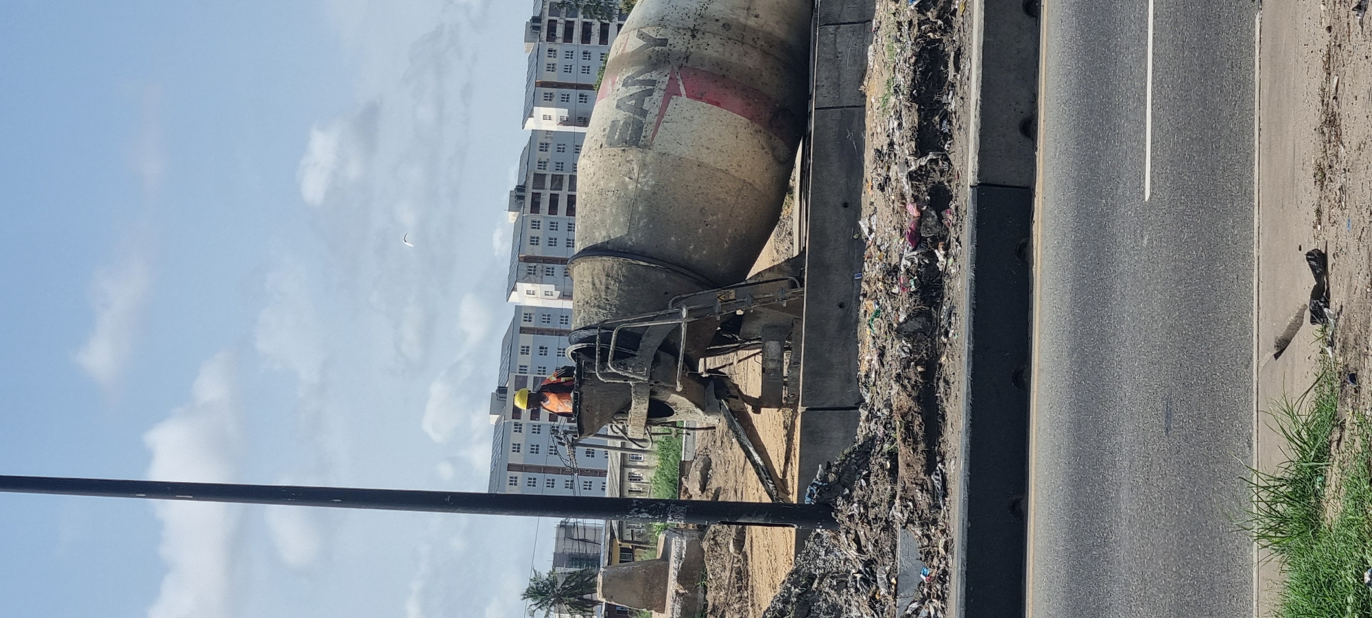 The image shows a construction site in Satellite Town, Nigeria. A large cement mixer truck is parked next to a road. A construction worker is visible near the mixer, wearing a yellow hard hat. The area around the mixer is littered with debris and trash. In the background, there are several residential buildings under construction. The sky is partly cloudy. A tall black pole is in the foreground. The road is asphalt with a white line marking. There is grass growing alongside the road.