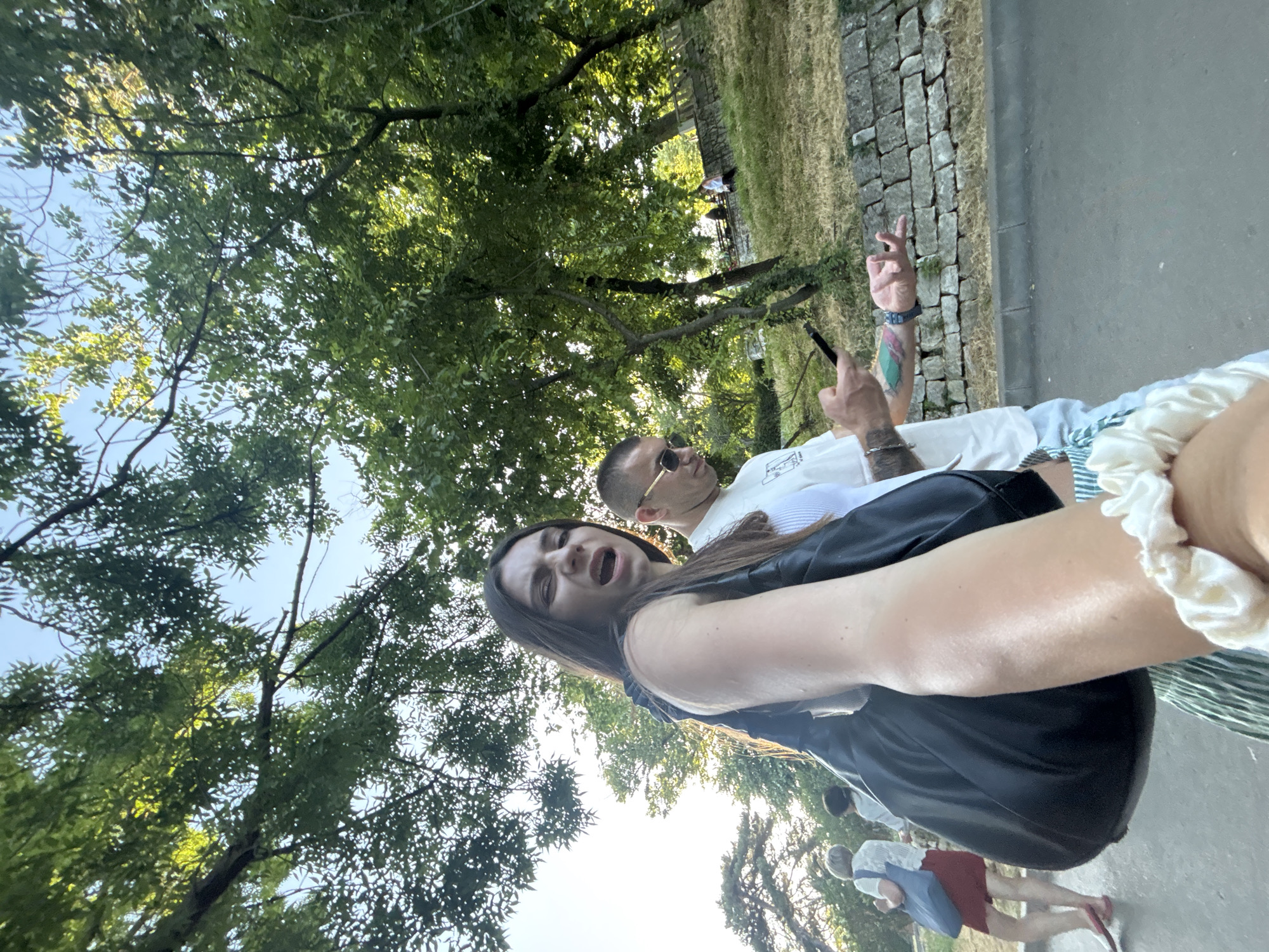 This is a portrait-oriented, low-angle, full-shot photograph taken outdoors on a sunny day. The foreground features a woman with long, dark hair and an open mouth, appearing to be looking at the camera and possibly speaking. She is wearing a black top. To her right and slightly behind her is a man wearing sunglasses and a white t-shirt, with tattoos visible on his arm. He is holding a phone and gesturing with his other hand. In the background, there are trees with green foliage, a stone wall, a grassy area, and a paved path. Two other people are visible in the background, one in a red skirt and another in a white shirt. The overall setting appears to be a park or outdoor public space, possibly Varna, Bulgaria, given the location context. The lighting suggests it is daytime, likely afternoon due to the warm tones. The woman's expression conveys surprise or excitement.