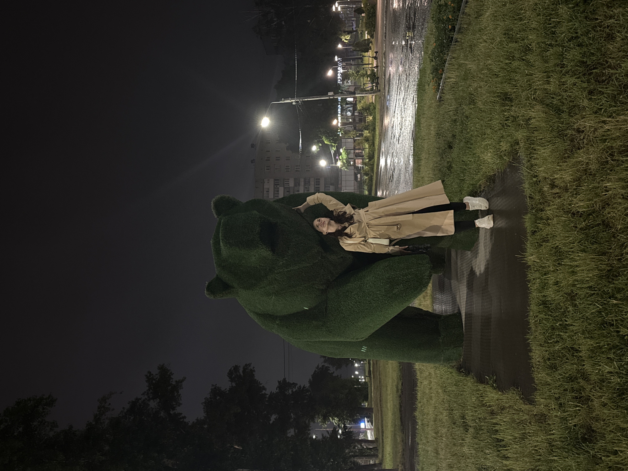 This nighttime photograph, taken in the city of Chaikovsky, Russia, features an adult female standing beside a large, green topiary sculpture of a bear. The woman, positioned on a wet paved pathway, is wearing a light beige trench coat, black trousers, and white sneakers. Her left arm is raised, with her hand touching the left side of the bear's head, and she is looking upwards towards her hand. The bear sculpture, covered in artificial green grass, is depicted in an upright, bipedal stance and significantly exceeds the woman's height. The ground surfaces, including the paved pathway and visible road, are wet and reflective, suggesting recent precipitation. The scene is illuminated by multiple artificial light sources, predominantly streetlights with bright white-yellow glows. In the background to the left, dark trees are visible, while to the right, across a wet street, multiple-story buildings and several parked electric scooters can be identified. The sky overhead is dark.