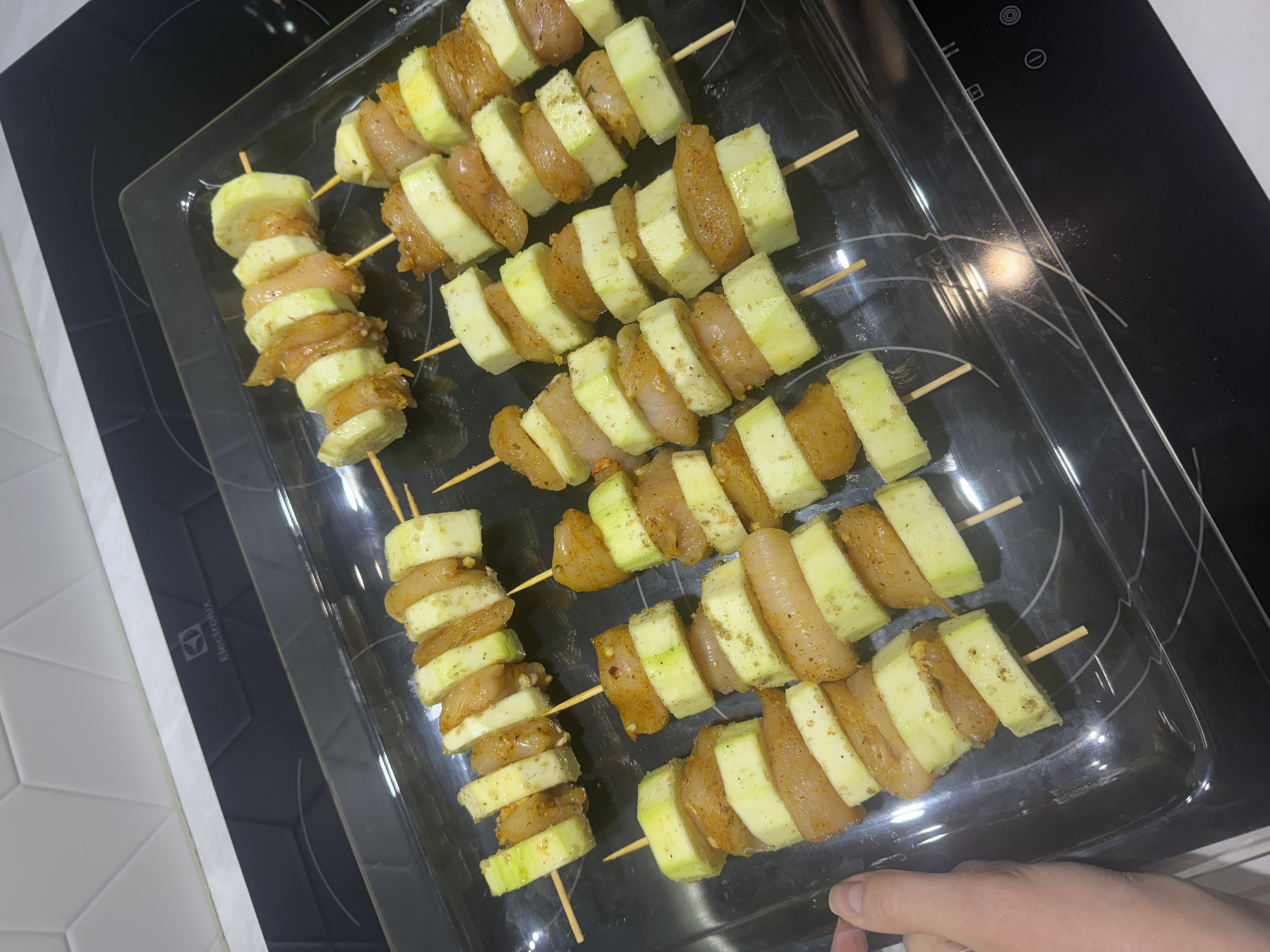 The image shows a clear rectangular glass baking dish containing several skewers of chicken and zucchini. The skewers are arranged in rows, with each skewer alternating between slices of zucchini and pieces of chicken. The chicken appears to be seasoned. The baking dish rests on a black glass cooktop. A hand is partially visible in the lower right corner of the image. The background includes a white tiled wall on the left. The branding "Electrolux" is visible on the cooktop. The image does not provide information relating to the city of Izhevsk.
