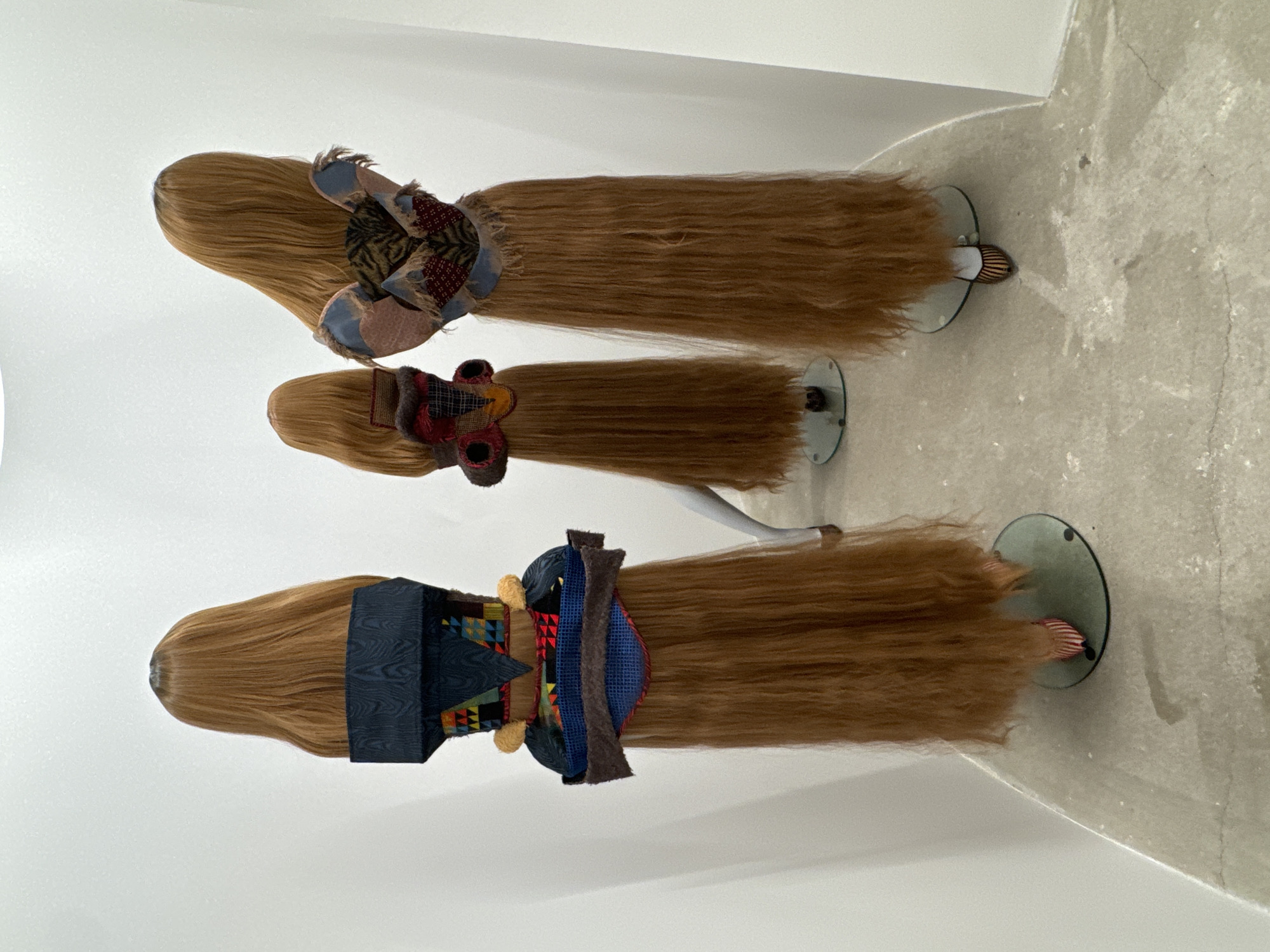 The image shows three stylized mannequin heads with long, straight, honey-brown hair, arranged in a row on a white, curved surface resembling a gallery display. Each mannequin wears a different type of fabric adornment or garment around the neck and shoulders. The mannequins are placed on clear, circular bases. The background consists of a white wall and a textured, light gray floor. The scene is likely indoors in a curated space. The location is unknown but the prompt suggested to mention that it is in Paris, France.