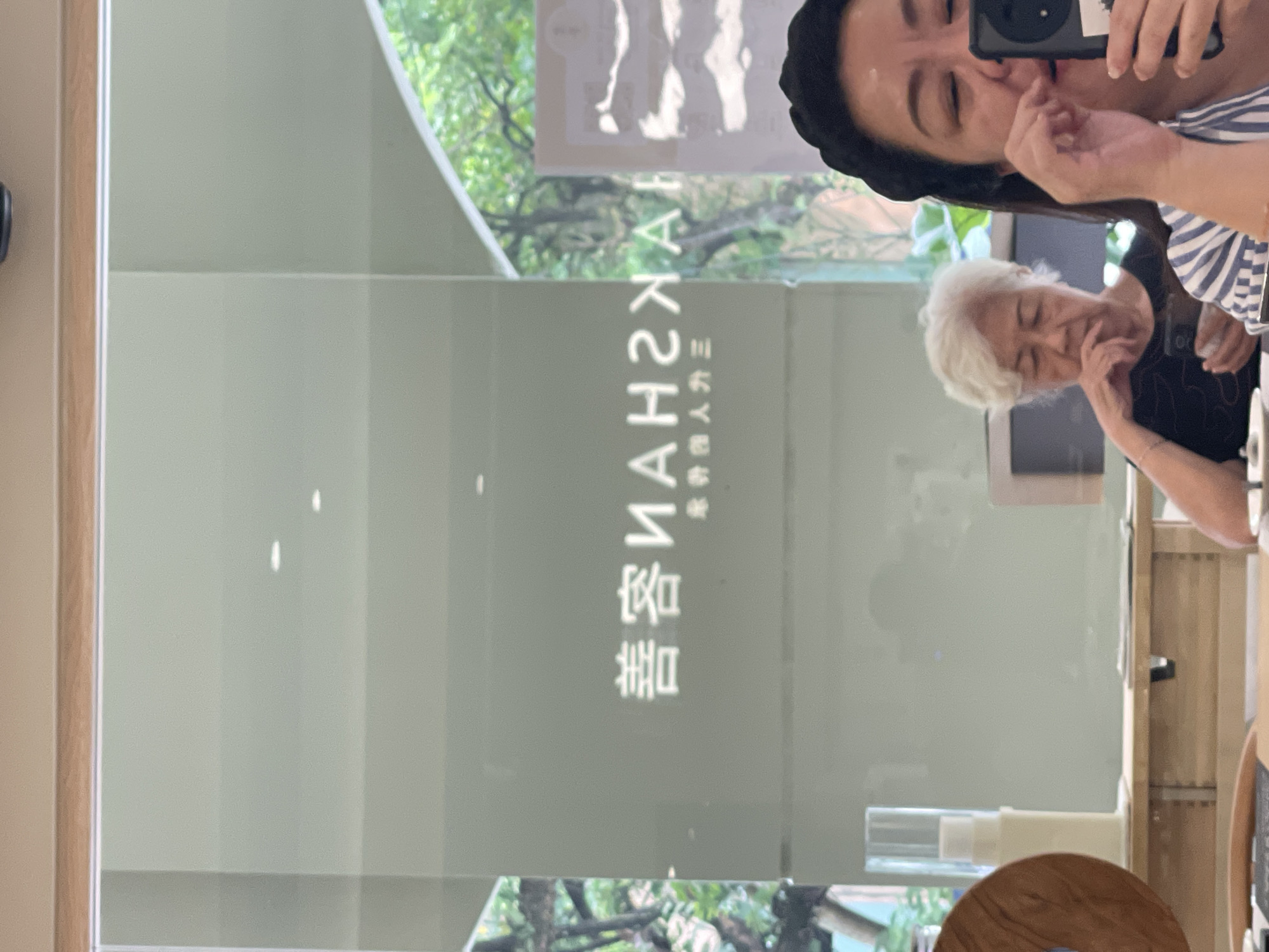 The image captures a reflection on a glass surface, revealing a scene in Kuala Lumpur, Malaysia. Prominently displayed on the glass are white characters, appearing to be in Chinese, with some reversed due to the reflection. Below these, smaller characters are also visible. In the foreground of the reflection, two individuals are present. On the right, a woman with dark hair, wearing a black headband and a blue and white striped shirt, is holding a black smartphone to her face, appearing to take a selfie or photograph. Her right hand is near her mouth. To her left and slightly further back, an elderly woman with white hair, wearing a black shirt, is looking down and holding a smartphone in her left hand. Both women are seated. Behind them, through the glass, a lush green outdoor environment with trees is visible. The overall setting appears to be an indoor establishment, possibly a cafe or restaurant, given the presence of tables and what looks like a counter area.