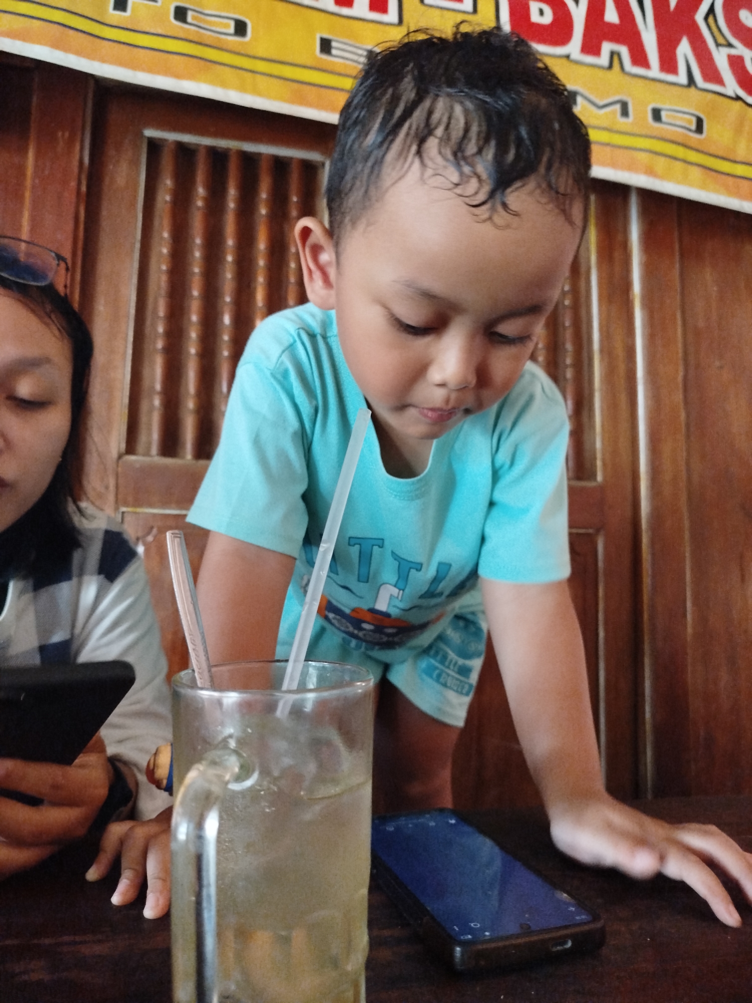 The image shows a young boy wearing a light blue t-shirt and shorts, leaning forward over a table. He has dark, curly hair and is looking down. To his left, a person with dark hair and glasses is looking down at a black smartphone in their left hand. In the center of the image, a glass of iced beverage with straws is visible. On the table in front of the boy is a dark-colored smartphone. The background consists of wooden paneling and a banner with text in red and yellow. The setting appears to be an indoor eating establishment.