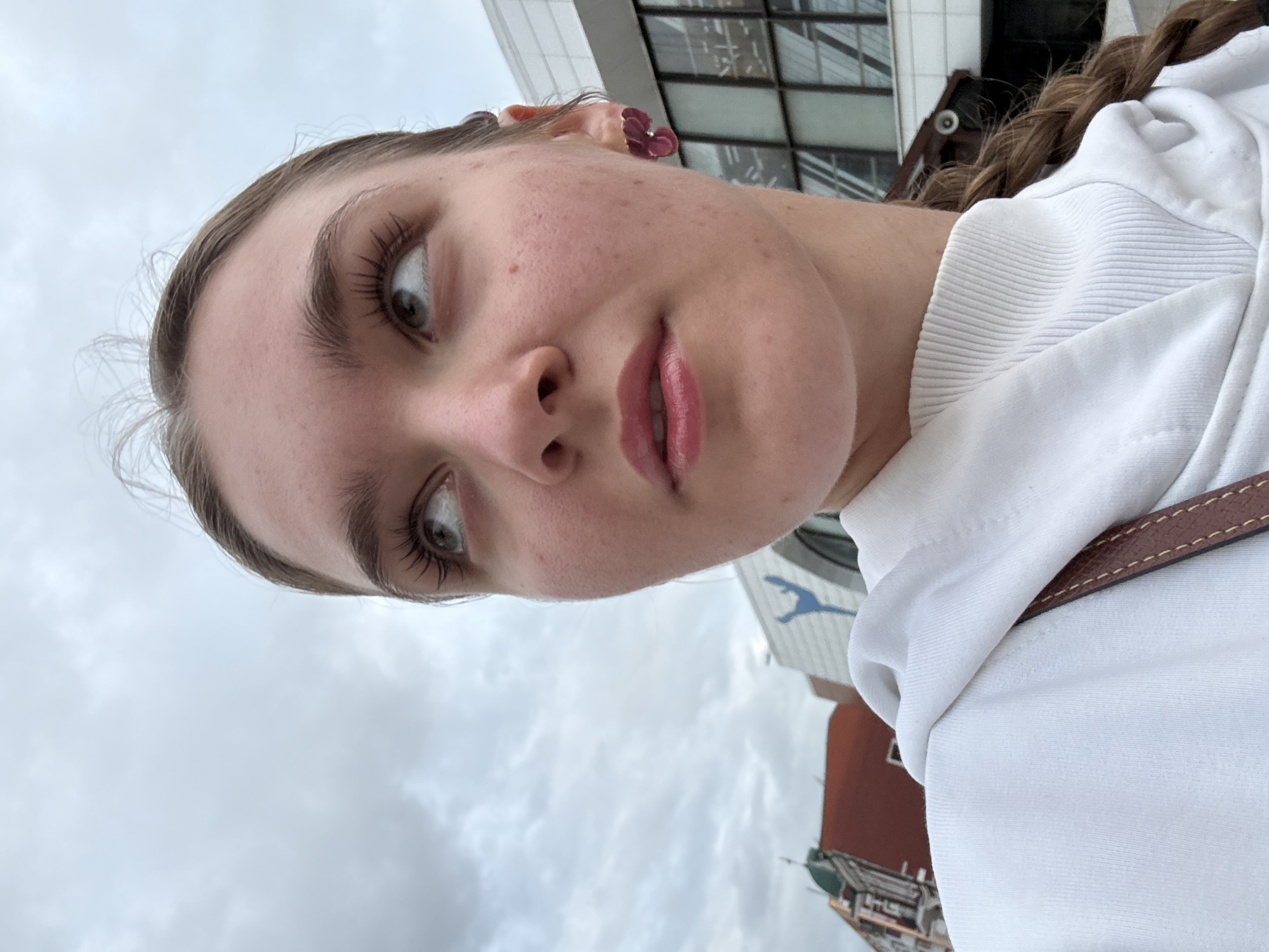 A low-angle, close-up photograph features a young female looking upwards and to the viewer's left. Her fair complexion shows slight redness on the cheeks, and her lips are glossy pink. She has long, dark eyelashes and light-colored eyes. Her light brown hair is styled in a braid, with some wisps visible at the hairline. She wears a white hooded sweatshirt over a white ribbed turtleneck. A reddish-purple, flower-shaped earring is visible in her right ear, and a brown leather strap from a bag rests on her left shoulder. The background consists of an overcast sky with grey clouds and urban architecture. To the left, part of an older building with a red tiled roof and a green dome is discernible. To the right, a modern building with a glass facade features a large, stylized blue graphic of a dynamic human figure, characteristic of the Perm Opera and Ballet Theatre in Perm, Russia.