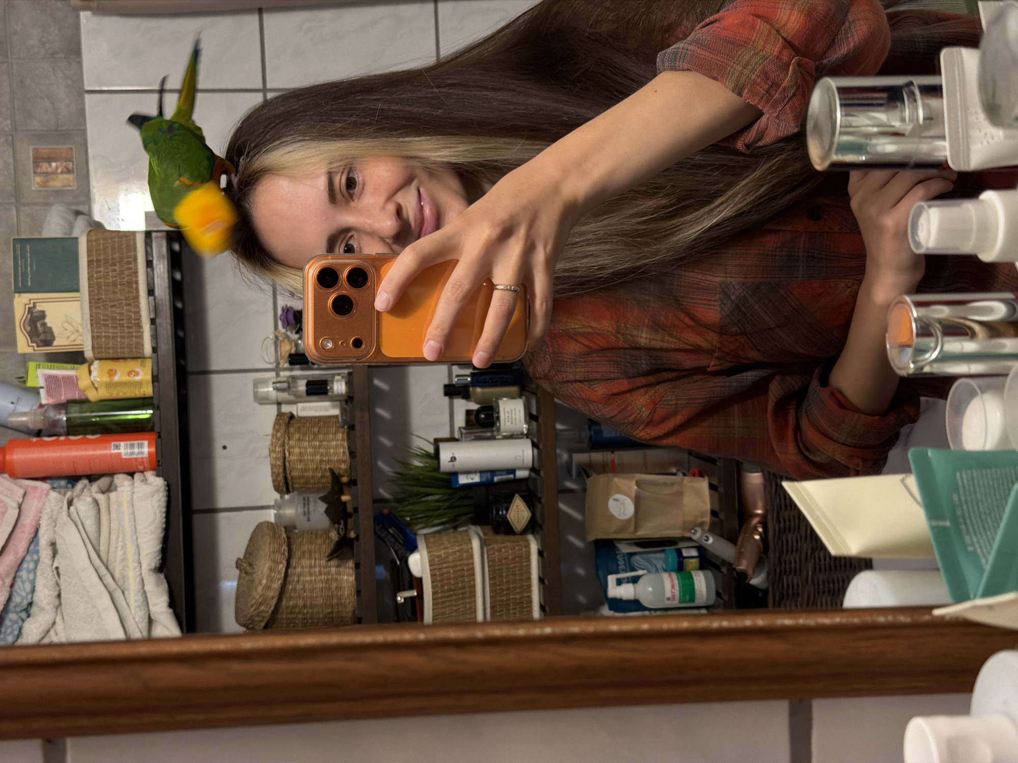 In a bathroom, a woman with long, brown hair, featuring blonde highlights, takes a selfie in a mirror. She is wearing a red and black plaid shirt. Perched on her head is a green and yellow parrot. The woman is holding an orange smartphone in her right hand, angled to capture the reflection. Her left hand is visible, with a ring on her ring finger. The bathroom shelves are stocked with various toiletries and decorative items, including woven baskets and bottles. The scene is captured from a medium shot, focusing on the woman and the parrot. The environment appears to be residential.