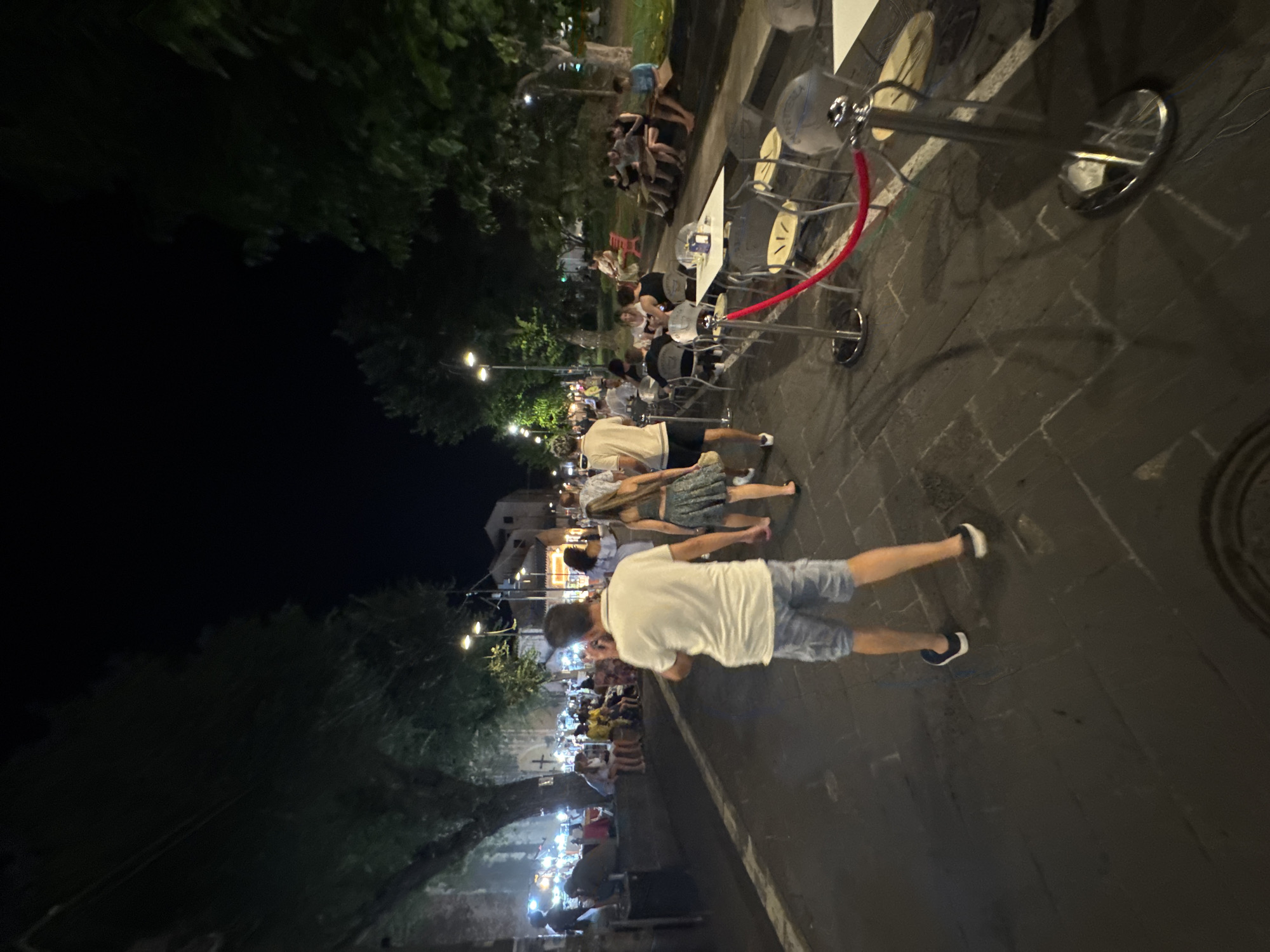 The image depicts a nighttime scene on a cobblestone street in Orosei, Italy. The street is lined with trees and buildings, with an outdoor seating area for a restaurant or cafe on the right side. Several people are visible, some walking and others seated at tables. The foreground shows the back of a man wearing a white polo shirt and blue shorts, walking away from the camera. To his right, a woman in a patterned skirt and top walks in the same direction. Further down the street, more people are gathered, some sitting at tables in the restaurant's outdoor seating area, and others standing or walking along the sidewalk. The lighting suggests it is evening, with streetlights and lights from buildings illuminating the scene. The overall atmosphere appears to be one of a pleasant summer evening, with people enjoying the outdoors.