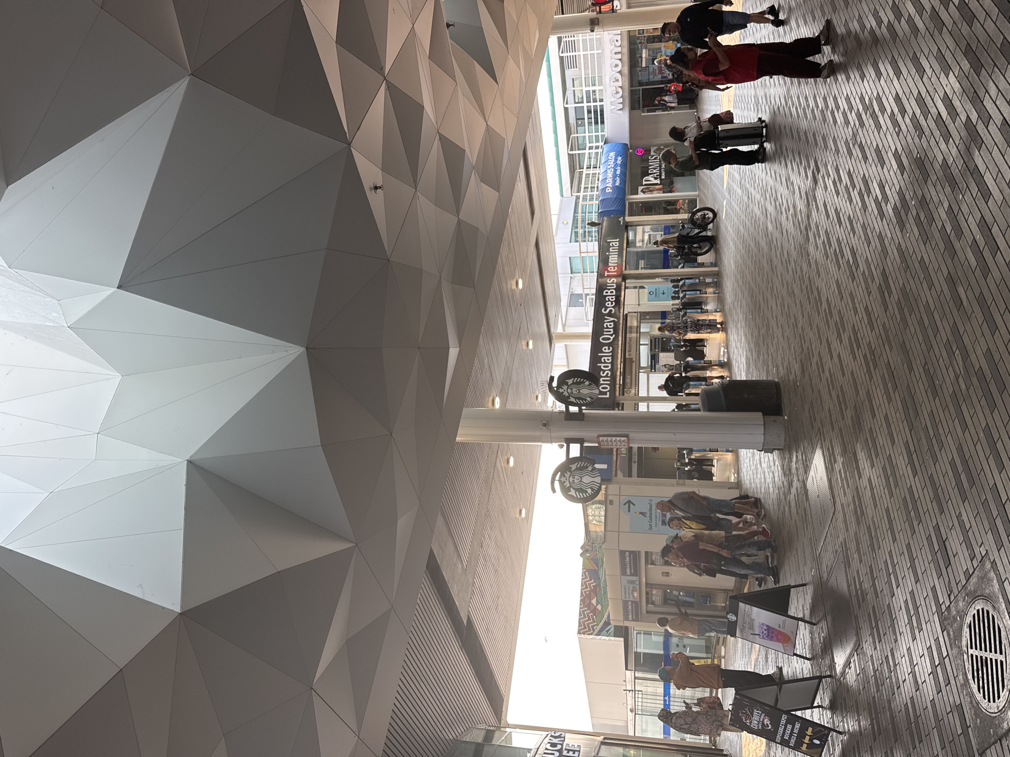 The image captures the entrance to the Lonsdale Quay SeaBus Terminal in North Vancouver, Canada, under a distinctive, modern, geometric-patterned canopy. Sunlight streams in from the right, illuminating the paved pedestrian area. A Starbucks sign is visible on the left, and further in, "Lonsdale Quay SeaBus Terminal" is displayed prominently. Several individuals are present, some queuing or walking. On the left, two A-frame signs are placed on the ground, one advertising "COWBOYS" and the other a Starbucks beverage. A McDonald's restaurant is also visible in the background on the right. The environment appears to be an outdoor public transit hub.