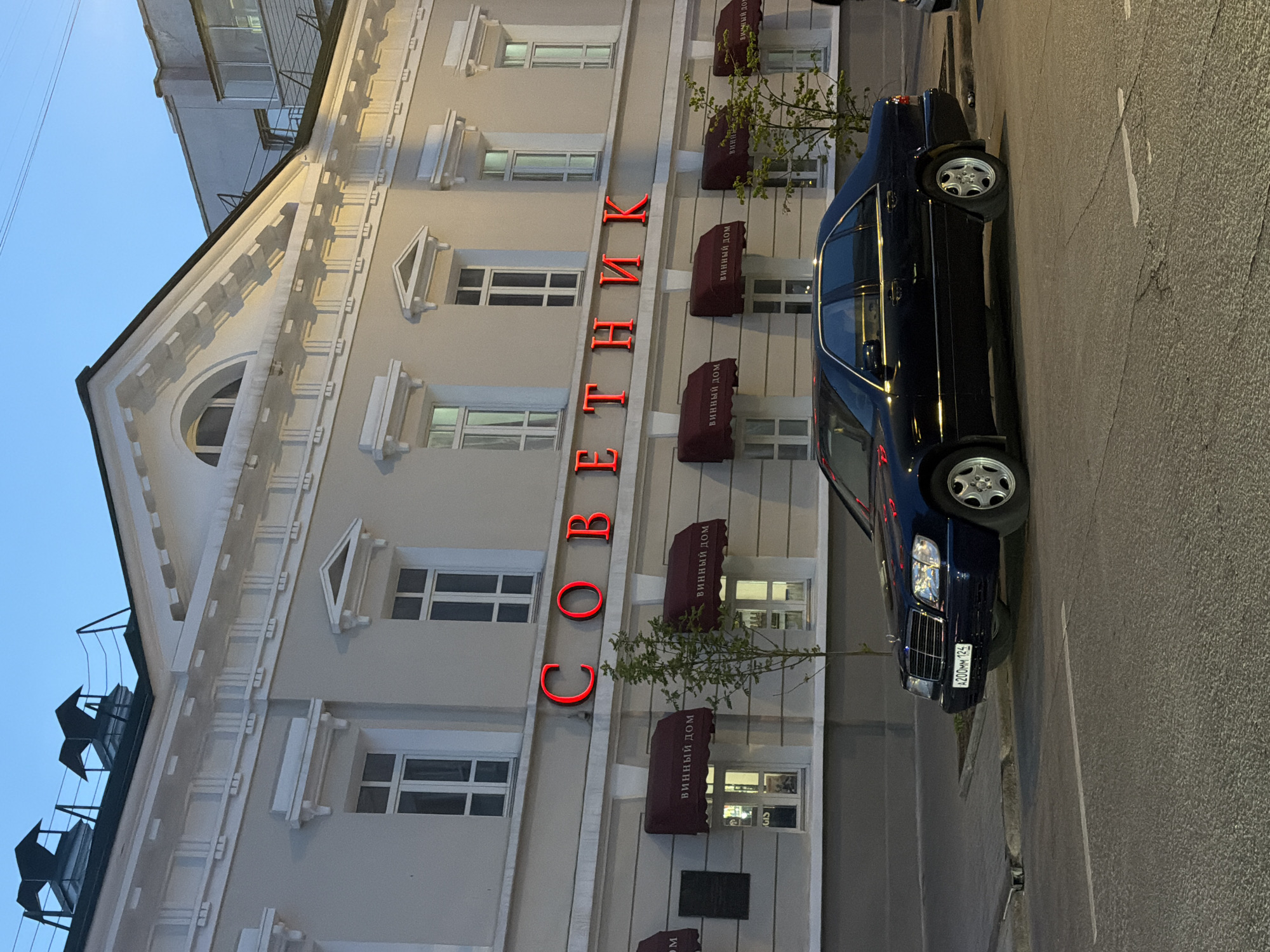 Here is a description of the image:
The image depicts a multi-story building in Krasnoyarsk, Russia. A black car, a Mercedes-Benz, is parked on the street in front of the building. The building has a gray facade with white architectural details. Vertically oriented neon red letters on the facade spell out "СОВЕТНИК." Below the lettering, there are awnings with the text "ВИННЫЙ ДОМ". Above the roof of the building are dark metal fixtures on the roof, while the sky is a dusky blue. The street appears to be paved with asphalt. The image is rotated.