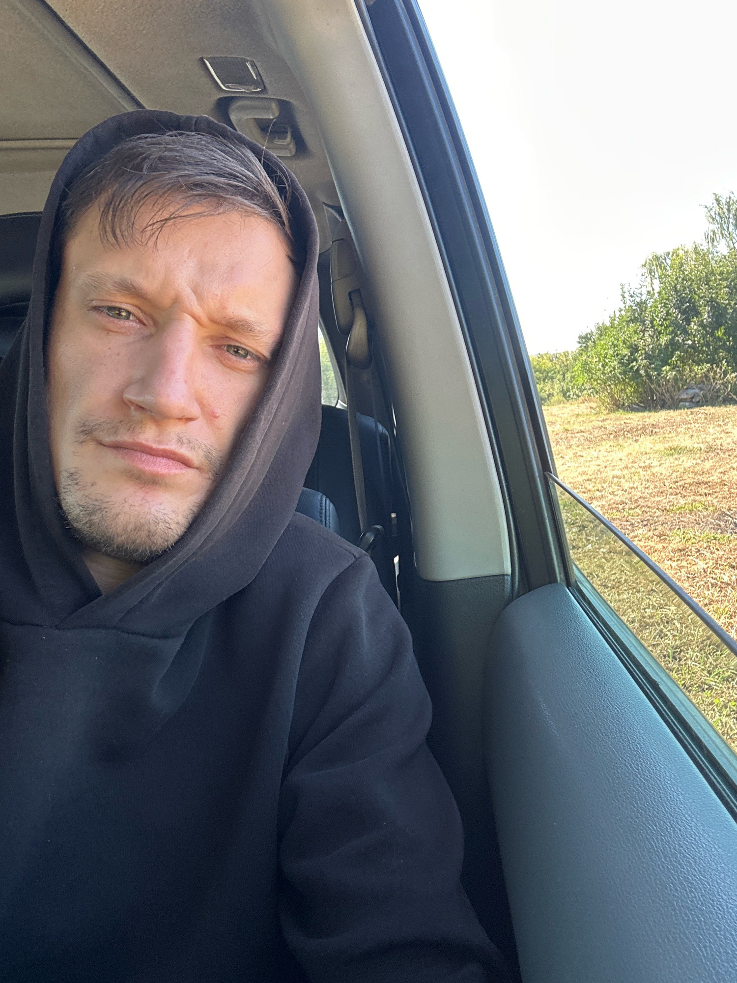 The image shows a close-up, selfie-style shot of a man wearing a black hoodie, with the hood pulled up over his head. He is inside a vehicle, indicated by the car interior visible in the foreground and the window on the right. The man is looking directly at the camera with a neutral, slightly furrowed expression. Beyond the car window, a dry, grassy field and green bushes are visible under a bright sky, suggesting an outdoor, possibly rural setting. The location in Makiv, Ukraine is not discernible from the image itself.