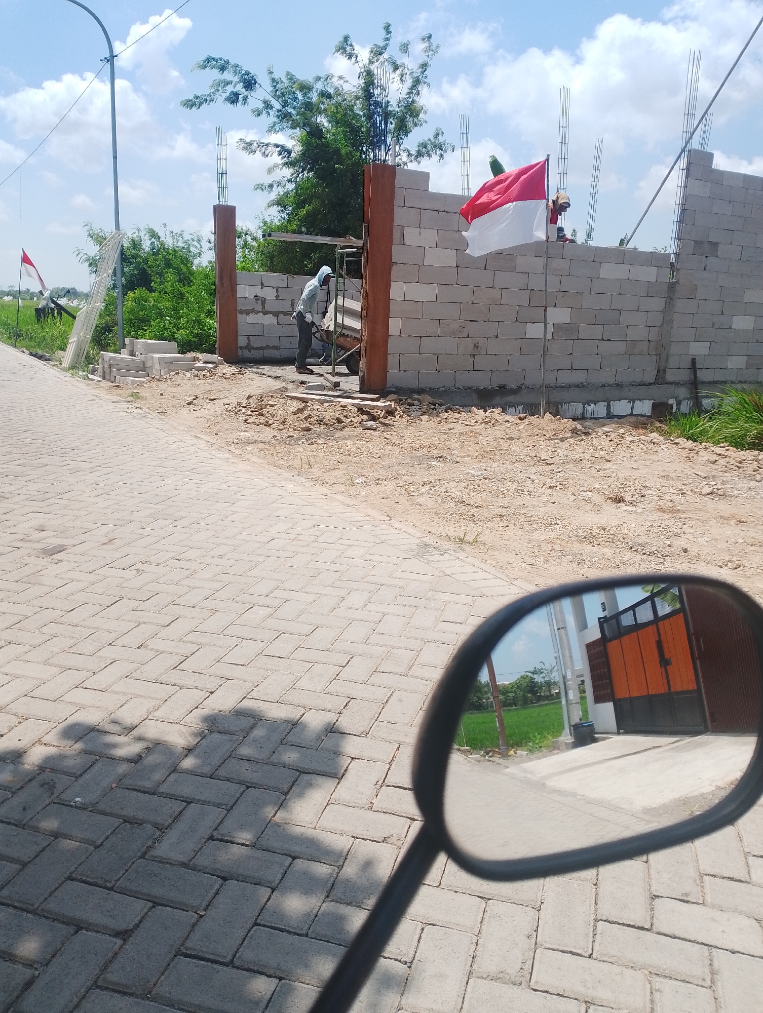 In Jumputrejo, Indonesia, a construction site is visible with a wall being built from grey cinder blocks. An Indonesian flag, red and white, flies from a pole in front of the wall. A worker wearing a grey hooded jacket and dark pants is pushing a wheelbarrow near the wall. A paved walkway, made of interlocking grey bricks, extends into the foreground. The reflection in a motorcycle's rearview mirror shows a bright orange gate with black trim and a glimpse of green rice fields. The environment is sunny with a blue sky and scattered white clouds.