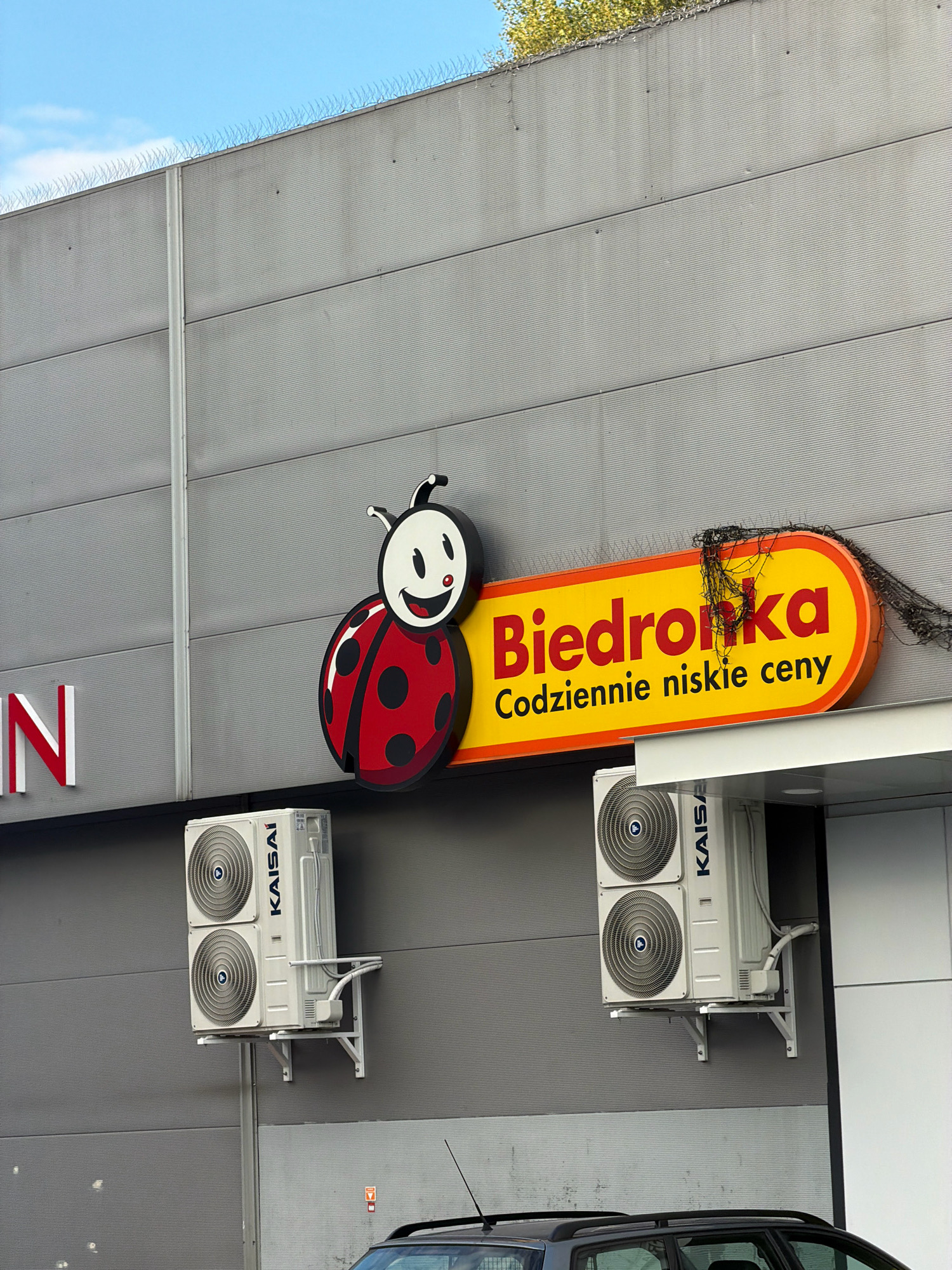 An outdoor shot shows the facade of a Biedronka supermarket in Wrocław, Poland. The building's exterior is a textured gray material. Prominently displayed is the Biedronka logo, a stylized ladybug with a smiling face, next to a bright yellow oval sign with red text that reads "Biedronka Codziennie niskie ceny" (Biedronka Daily low prices). Two white outdoor air conditioning units are mounted on the wall below the sign. To the left of the logo, partially visible, is a white sign with red letters that appears to say "IN". The sky above is blue with some clouds, and on the top edge of the building, anti-bird spikes are visible. Below the facade, the roof of a dark-colored car is visible.