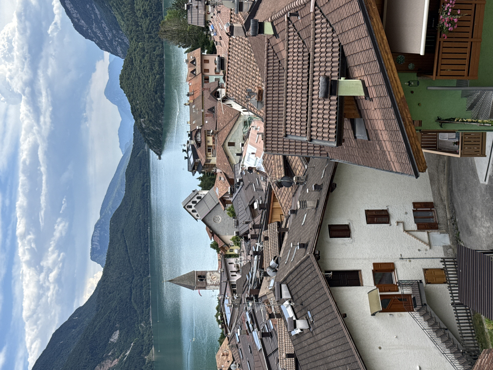 The image shows a panoramic view of the town of Molveno, Italy, nestled beside a lake and surrounded by mountains. In the foreground, numerous terracotta-tiled roofs of houses dominate the scene, with various chimneys and satellite dishes visible. A white stucco house with a dark-tiled roof and a steep outdoor staircase leading to an entrance is prominent on the left side of the frame. Further into the midground, other buildings with similar architectural styles and tiled roofs are visible, including a church with a tall, pointed steeple and a clock face. The lake, a vibrant turquoise color, stretches across the background, reflecting the sky. The distant mountains are covered in lush green forests, with a few rocky peaks visible in the hazy distance. The sky is a mix of blue and white clouds. There are no people visible in the image, but small boats are scattered on the lake, and a paraglider is visible in the sky.