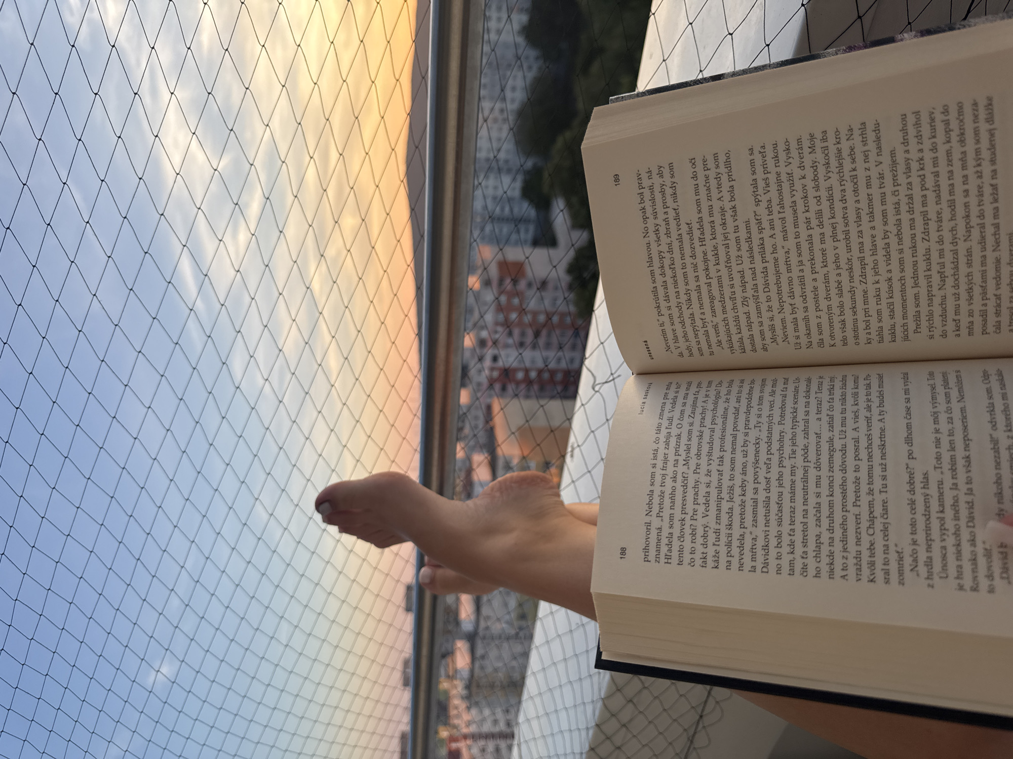 In Bratislava, Slovakia, a pair of bare feet rests on a book, positioned in front of a balcony railing. The book, open to pages 188 and 189, is partially obscured by the feet. Beyond the balcony, the cityscape unfolds with buildings and trees, under a soft gradient sky transitioning from blue to orange. A fine mesh netting is visible in the foreground, creating a diamond pattern that overlays the scene. The lighting suggests it is either sunrise or sunset.