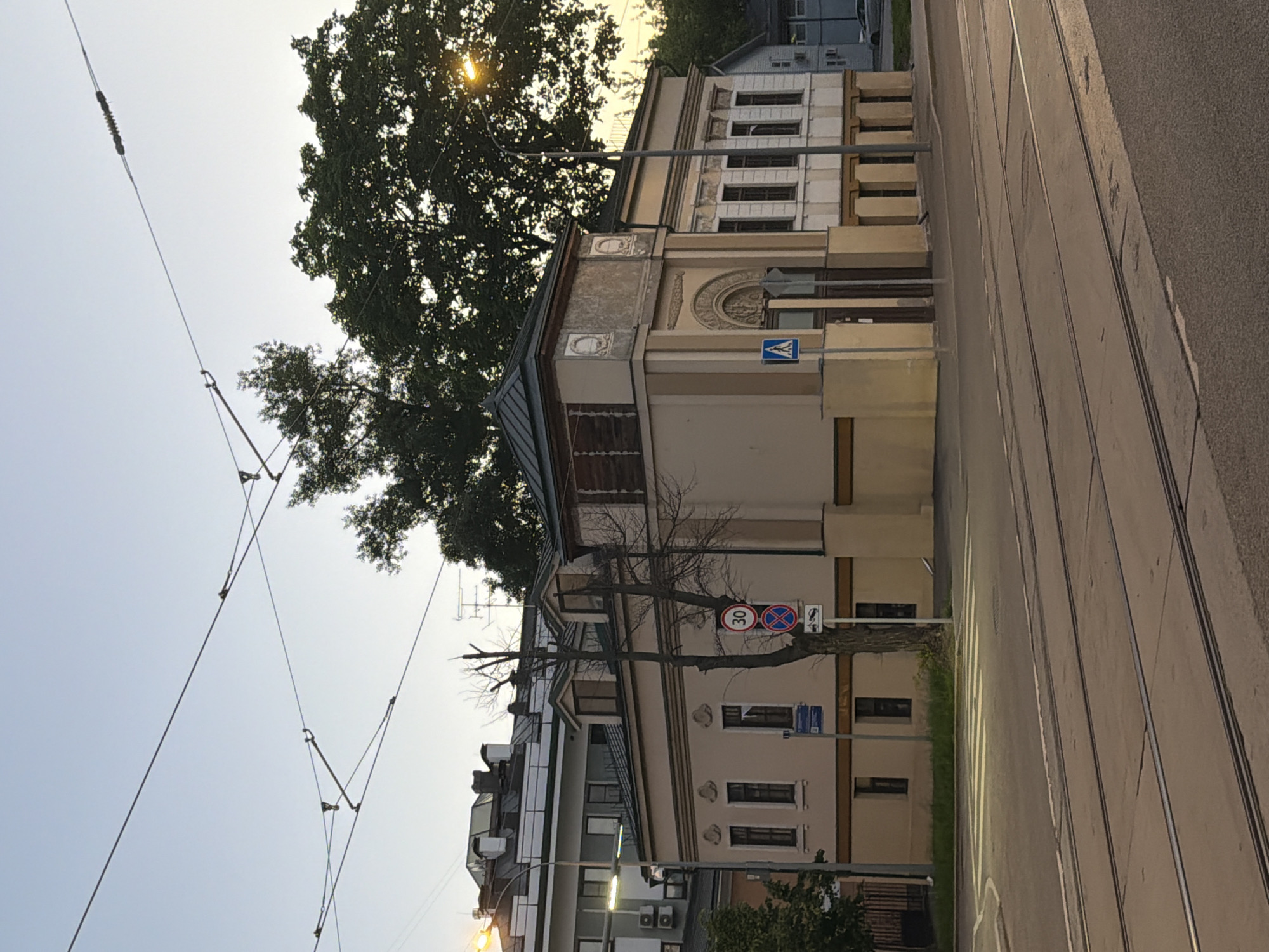 The image depicts a street scene in Moscow, Russia, featuring a beige building with ornate architectural details and windows along the side of the road. A large tree partially obscures the upper portion of the building, casting shadows on its facade. Overhead tram lines and wires stretch across the sky, intersecting with the trees. The road has tram tracks and street markings. Road signs are visible, indicating traffic regulations. A pedestrian crossing sign is also present. The sky is pale, suggesting either dawn or dusk. Other buildings and street lights are visible in the background, along with smaller trees and greenery.