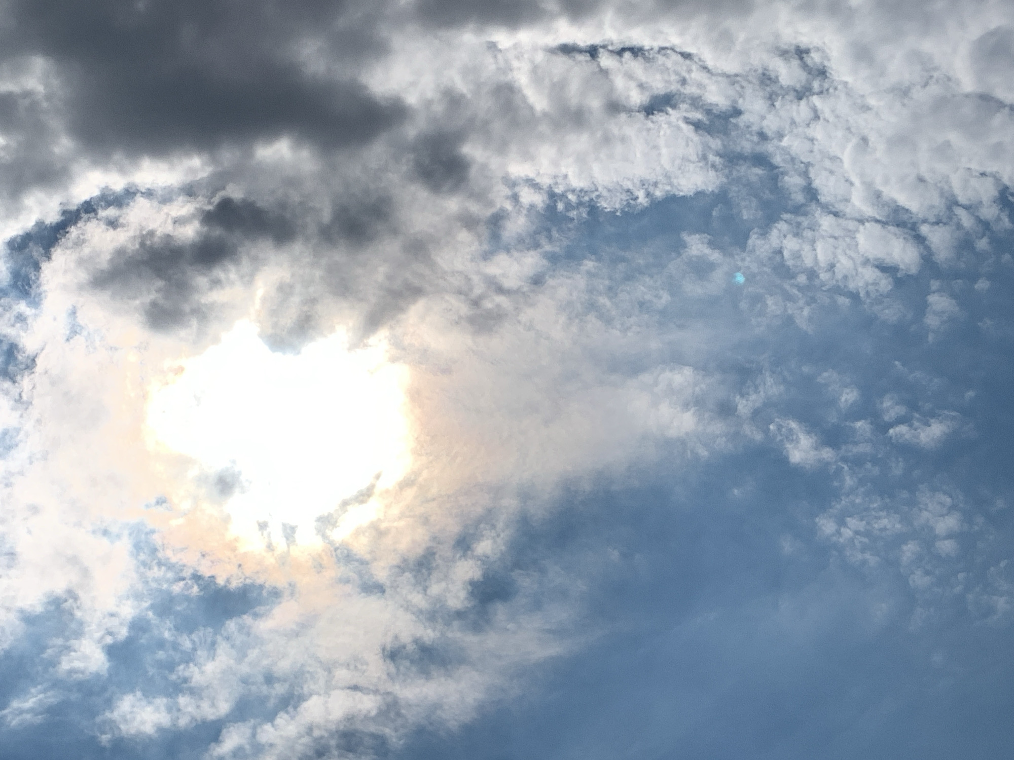 A wide-angle, low-angle shot captures a dynamic sky above Khazar Raion, Azerbaijan. The sky is filled with a mix of dark, heavy clouds on the left and lighter, scattered clouds on the right, creating a dramatic contrast. The sun is partially obscured by the clouds, casting a bright, diffused light that creates a luminous halo effect. The light appears to have a warm, slightly pinkish hue, suggesting either a sunrise or sunset. The lower portion of the image transitions into a clear blue sky, with some wispy clouds visible. There are no people, structures, or terrestrial objects in the frame, focusing solely on the atmospheric conditions of the sky. The overall impression is one of natural beauty and the power of the elements.