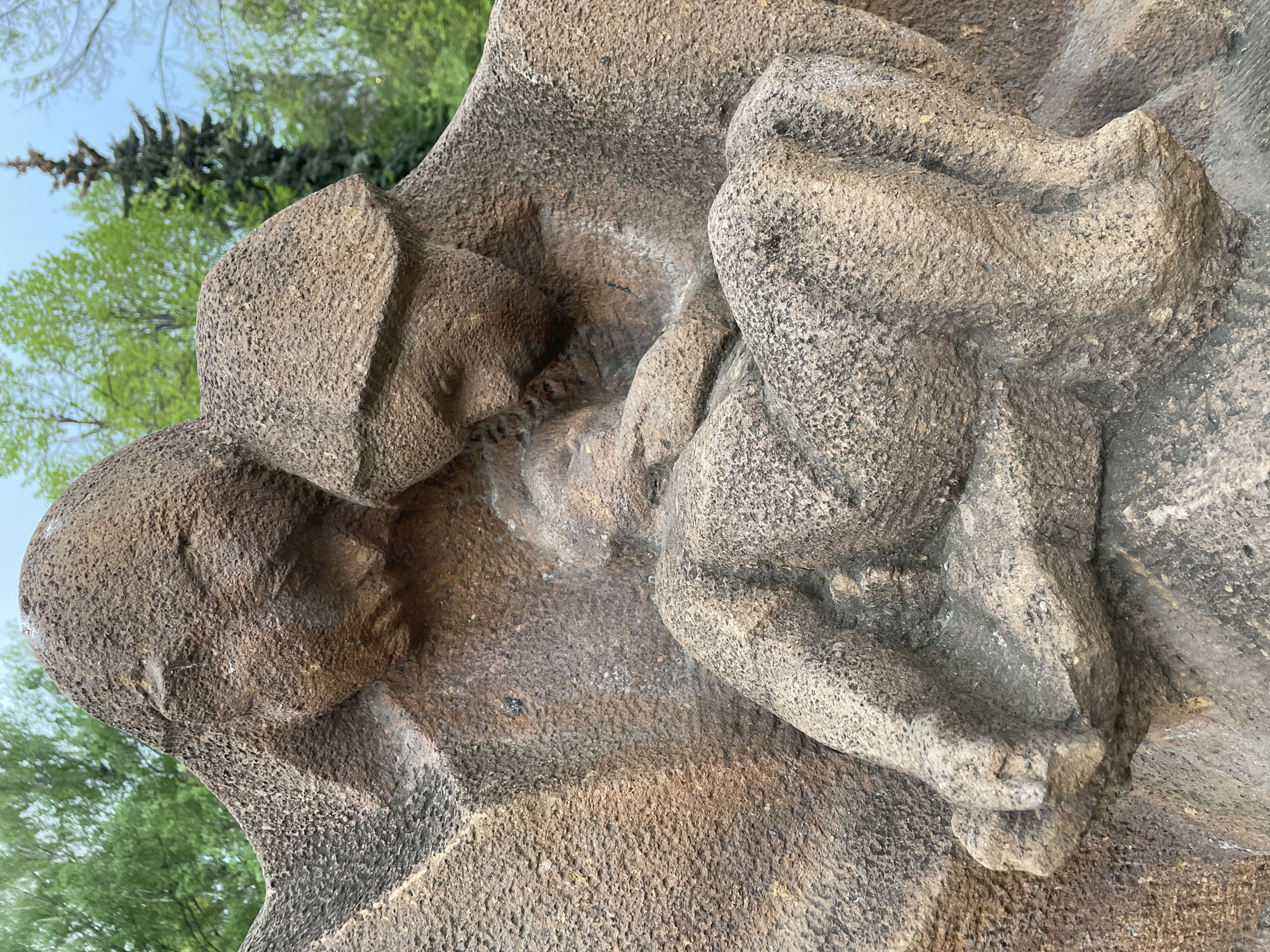 A close-up, tilted, color photograph shows a stone sculpture of two figures. The figure on the left is larger and appears to be supporting a smaller, curled-up figure on the right. Both figures are rendered in a rough, textured stone. Green trees and a light blue sky are visible in the background. Based on location clues, the image depicts a section of a monument likely situated in Khimki, Russia.