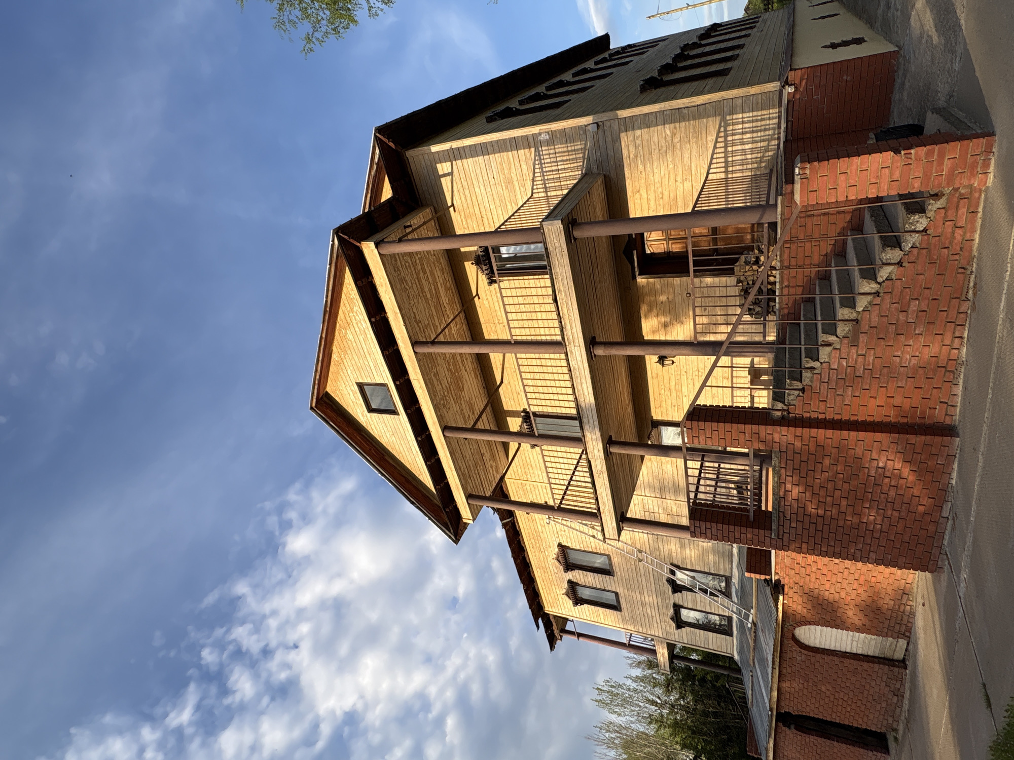 A wooden house with brick accents is the focal point. The house features balconies on the middle two stories, supported by metal columns. The roof has a steep pitch with a small dormer window near the top. The sky is blue with scattered clouds. The house has a staircase leading up to a side entrance on the first floor. The photograph was taken in Zavod Mikhailovskii, Russia.