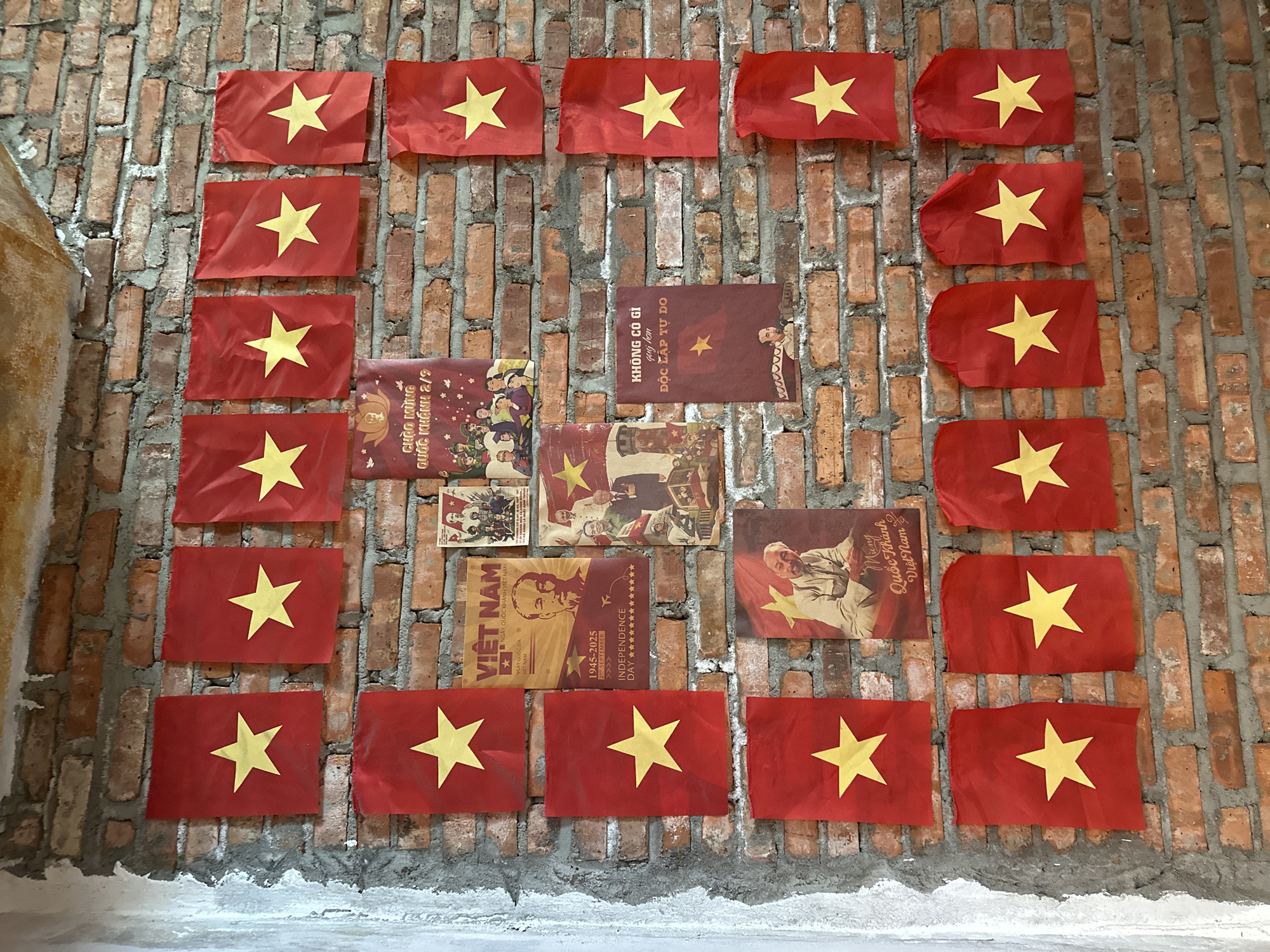 The image displays a brick wall adorned with numerous small Vietnamese flags and propaganda posters, located in Đà Nẵng, Vietnam. The wall is constructed from reddish-brown bricks with grey mortar, exhibiting some discoloration and rough plaster on the far left edge. Arranged in a grid pattern across the wall are approximately 20-25 rectangular red flags, each featuring a central yellow five-pointed star. These flags appear to be made of fabric and are attached directly to the wall, showing natural creases and folds.
Interspersed among the flags are several vintage-style posters. A red poster in the top center depicts a group of figures and bears the Vietnamese text "CHÀO MỪNG QUỐC KHÁNH 2/9" (Welcome National Day 2/9). Below this, a prominent yellow-orange poster features a portrait of Ho Chi Minh, with the text "VIỆT NAM NGÀY QUỐC KHÁNH VIỆT NAM" and "1945-2025 2nd SEPTEMBER INDEPENDENCE DAY." To its right, another poster includes a group of soldiers or figures and text containing the phrase "KHÔNG CÓ GÌ quý hơn ĐỘC LẬP TỰ DO" (Nothing is more precious than independence and freedom), attributed to Ho Chi Minh. A lower-left poster shows a smiling Ho Chi Minh, with text likely relating to Vietnam's National Day. The arrangement forms a thematic display celebrating Vietnamese national identity and independence, specifically referencing September 2nd.