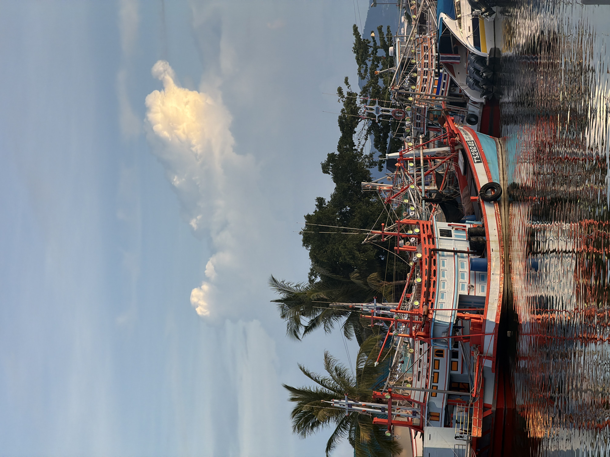 In the city of ตำบลเกาะพะงัน, Thailand, a fleet of colorful fishing boats is docked in a harbor under a sky filled with soft, diffused sunlight and scattered clouds. The boats, primarily painted in red and white with blue accents, are intricately rigged with numerous poles and lines, suggesting their readiness for fishing. The foreground is dominated by the calm water reflecting the vibrant colors and structures of the vessels, creating a mirror-like effect. Palm trees and dense green foliage are visible behind the boats, bordering the shoreline and hinting at a tropical coastal environment. The scene captures a moment of stillness before or after a day's work at sea, with the setting or rising sun casting a warm glow on the water and the prominent, brightly lit cloud formation in the upper right quadrant of the sky.