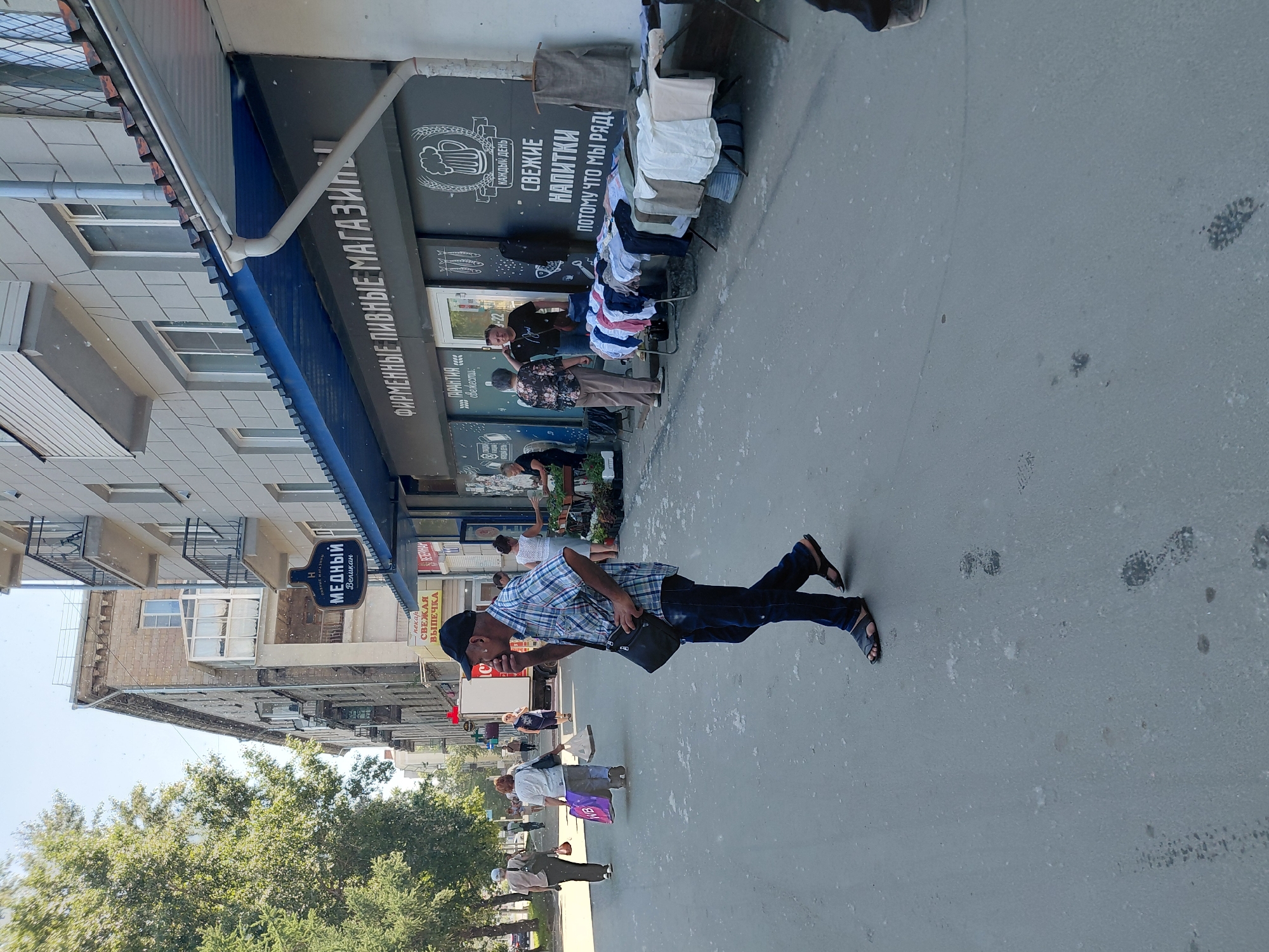 A man in a checkered shirt and dark pants walks on a paved sidewalk next to a building with shops. He is wearing sandals and carrying a black messenger bag. In front of the building, tables are set up displaying various clothing items. Several other people are visible in the background, including shoppers and people entering and exiting the shops. The building has a dark facade with text in Russian, including "ФИРМЕННЫЕ ПИВНЫЕ МАГАЗИНЫ" which translates to "Brand Beer Stores." Other signs indicate "СВЕЖИЕ НАПИТКИ" or "Fresh Drinks." The environment appears to be an urban setting in Novosibirsk, Russia, with trees lining the street and a clear sky overhead. The ground is asphalt and shows some scattered debris.