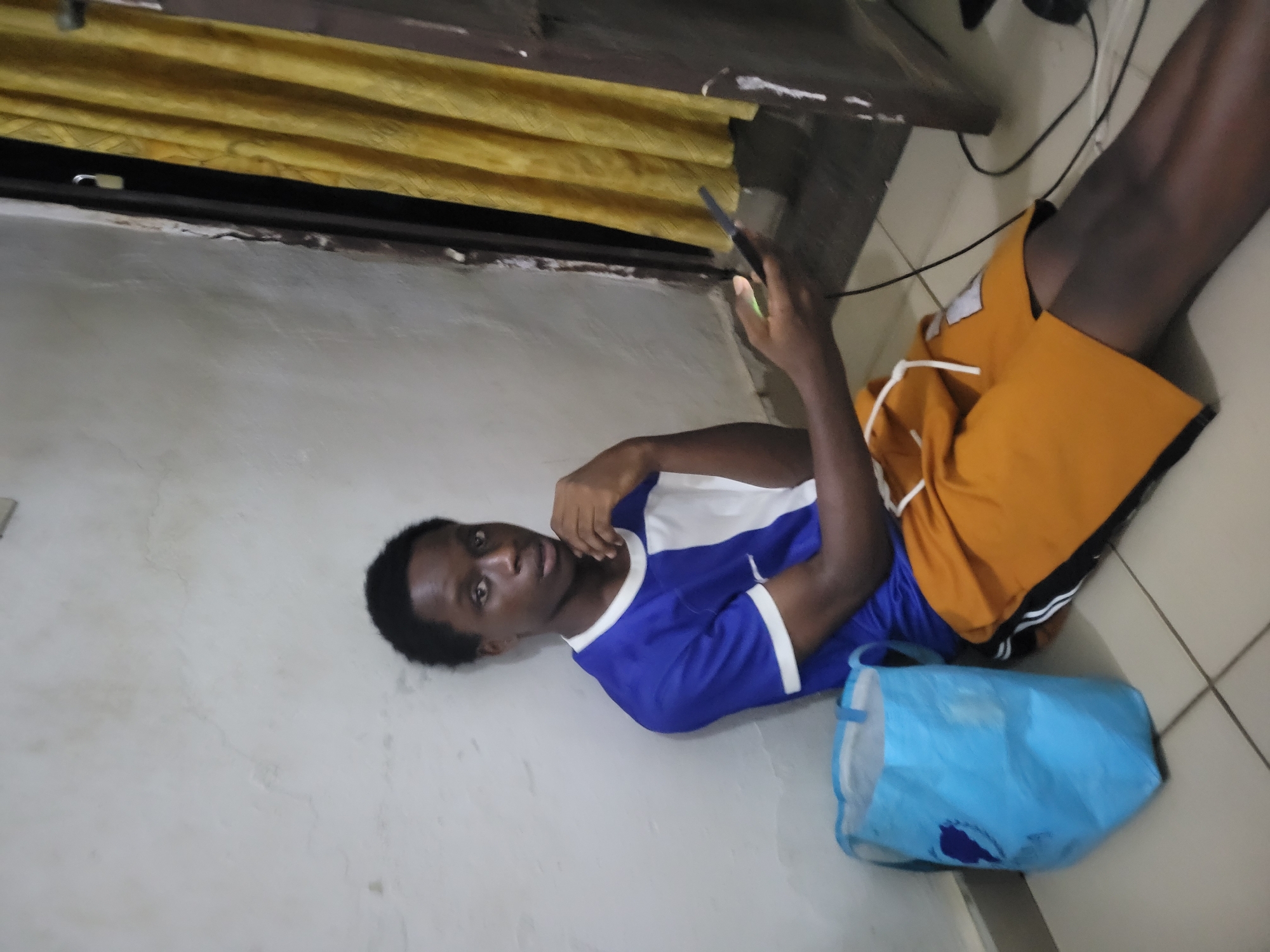 A young Black male is seated on the floor, leaning against a textured off-white wall, in what appears to be an indoor setting. He is wearing a blue and white t-shirt and orange shorts. His left hand is resting on his chin, and his right hand is holding a black mobile phone, with a charging cable visible connecting to it. Beside him, to his left, is a light blue tote bag. The background features a yellow curtain partially covering a doorway or window. The floor is tiled. The lighting suggests it is likely evening or indoors with artificial lighting.
