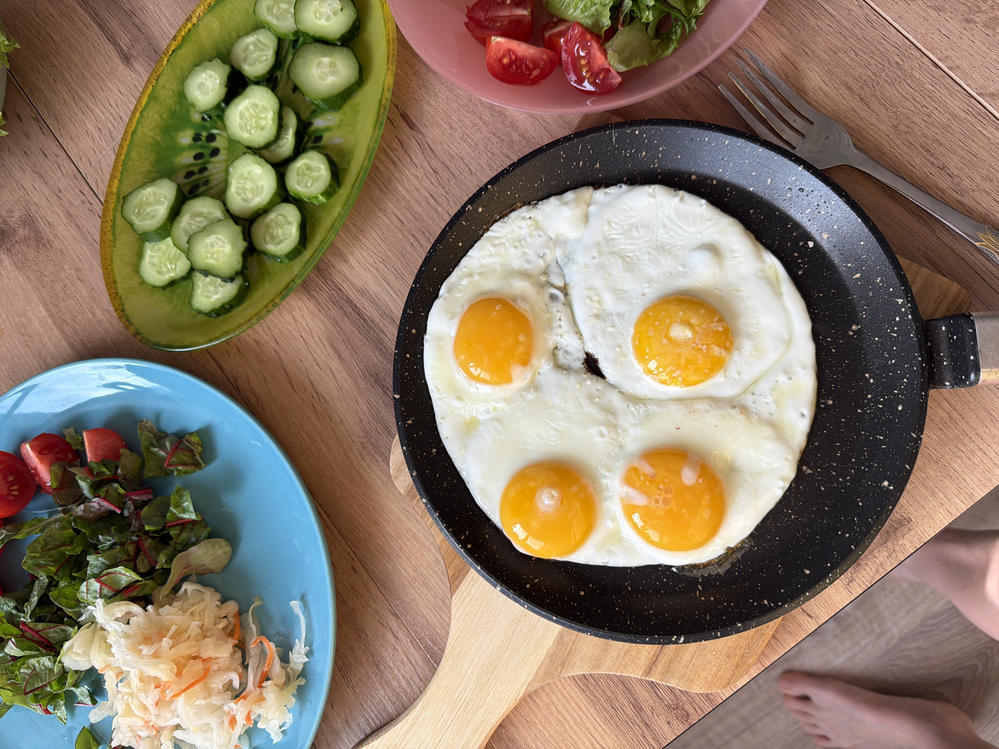 The image depicts a breakfast setting on a wooden table. A black frying pan with a light-colored handle contains four sunny-side-up eggs. Adjacent to the pan, there's a fork resting on the table. Three plates surround the pan: a green plate with sliced cucumbers, a pink plate with tomatoes and lettuce, and a blue plate with greens, tomatoes, and what appears to be sauerkraut. The background shows part of a person's foot on a light-colored surface. There's no information about this image's location being in Izhevsk, Russia.