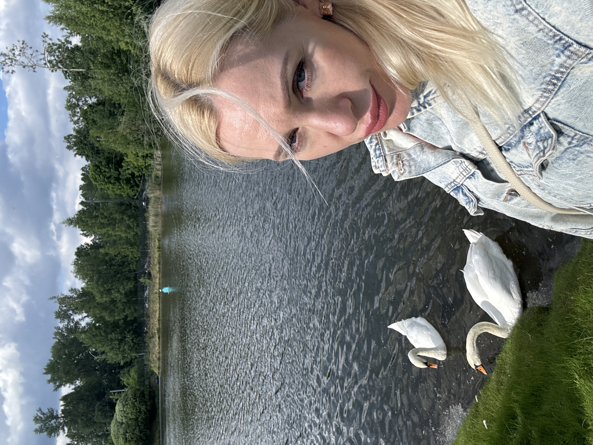 The original image is a selfie taken by a blonde woman in Saint Petersburg, Russia. She is wearing a denim jacket and standing near a body of water with two swans swimming. The background features trees and a cloudy sky. The location appears to be a park or natural area within the city.